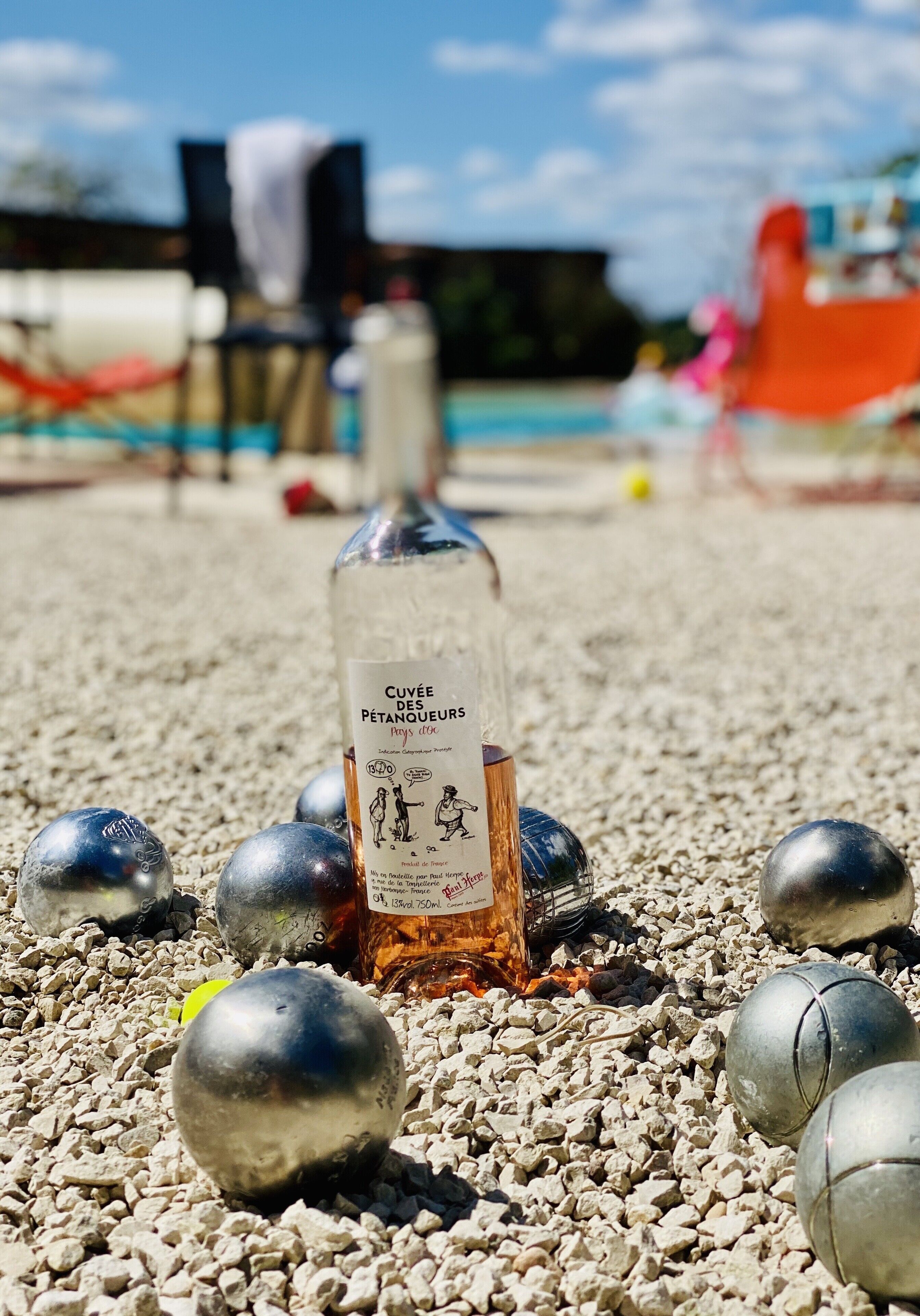 Una botella de Cuvée des Pétanques sobre un terreno de grava con bolas de billar alrededor.