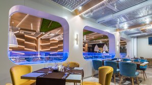 Een modern restaurant met een opvallend geometrisch plafond, grote ramen en een combinatie van gele en blauwe stoelen.