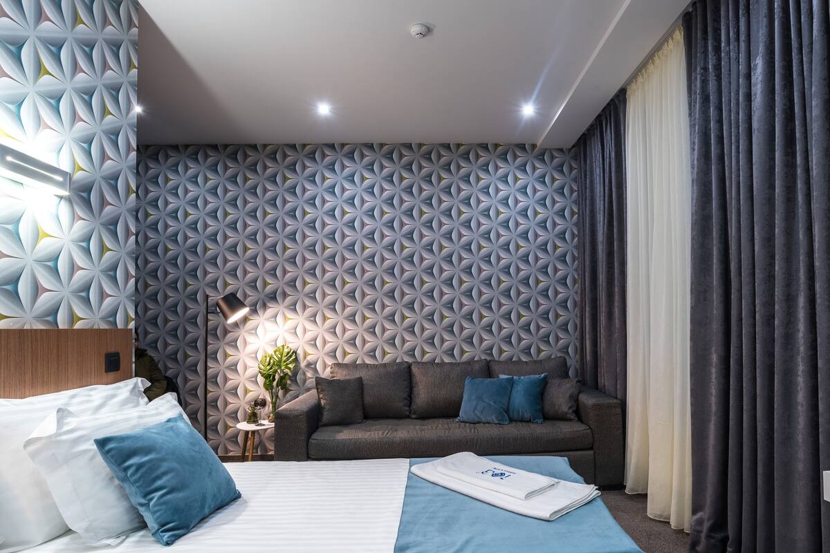 A modern hotel room with a patterned wall, a sofa, a bed, and a desk.