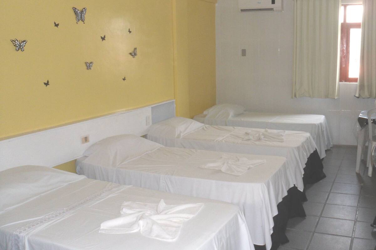 A hotel room with three single beds, a yellow wall, butterfly wall decals, an air conditioning unit, and a window with curtains.