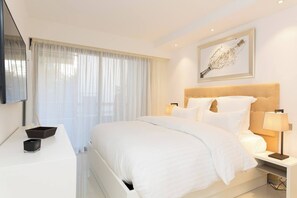 Quarto de hotel com uma cama grande, mesa de cabeceira com abajur, televisão na parede e janela com cortinas leves.