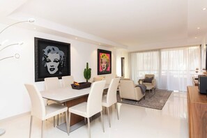 A modern living room with a dining area, featuring a flat-screen TV, a large window with sheer curtains, and artwork on the walls.