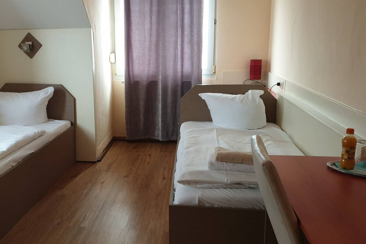 A hotel room with two single beds, a wooden floor, a small table with a bottle, and a window with curtains.
