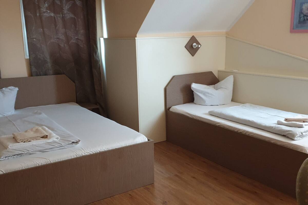 A hotel room with two beds, one larger and one smaller, both with white linens. There is a chair and a small table in the corner.