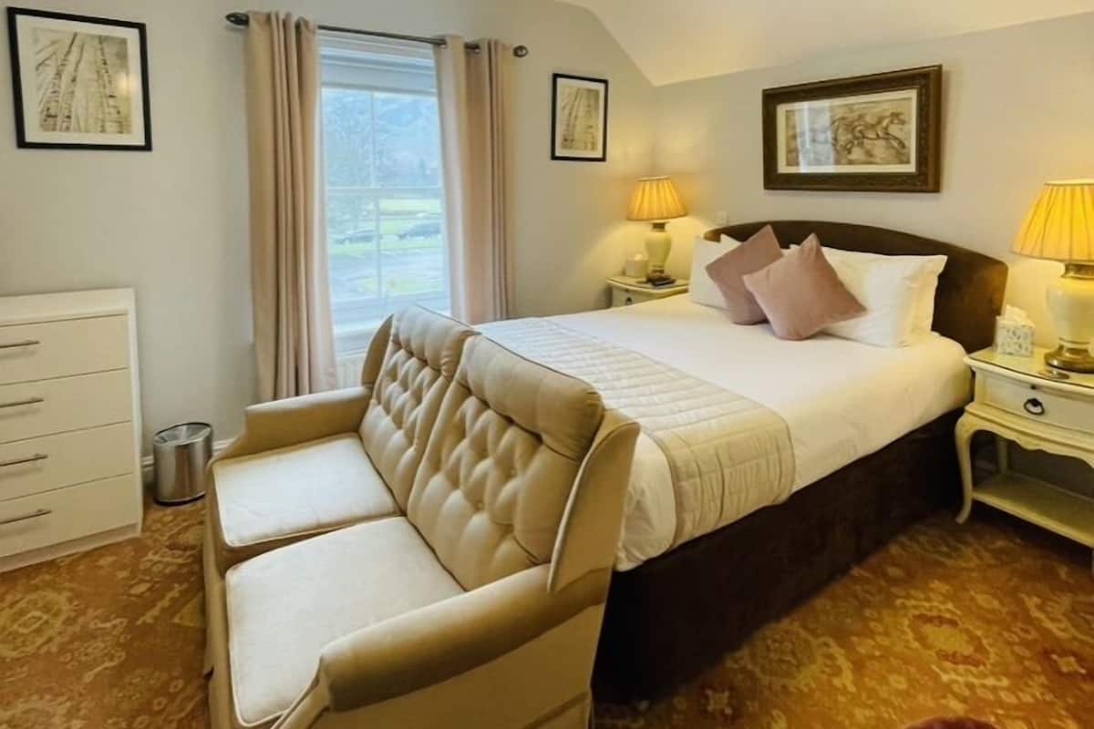 A hotel room with a bed, a sofa, a nightstand with a lamp, a window with curtains, and framed pictures on the walls.
