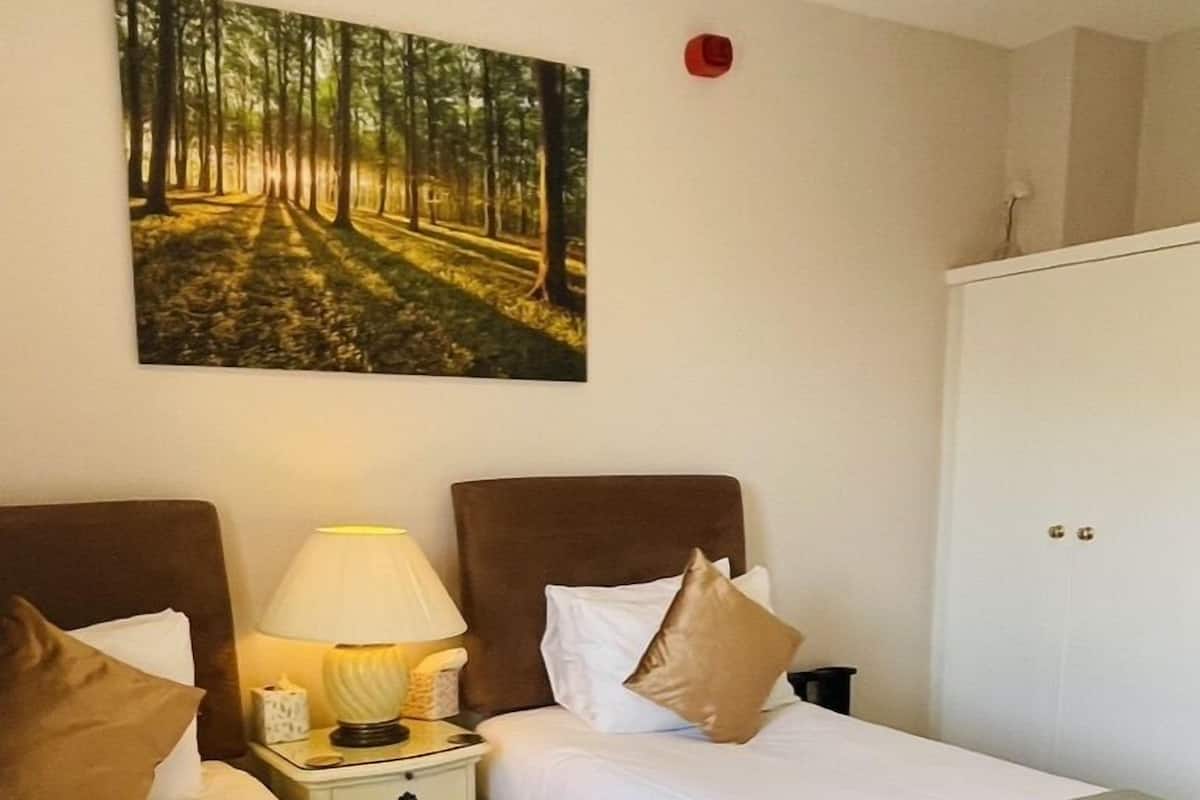 A hotel room with two beds, a nightstand with a lamp, a painting of a forest, and a wardrobe.