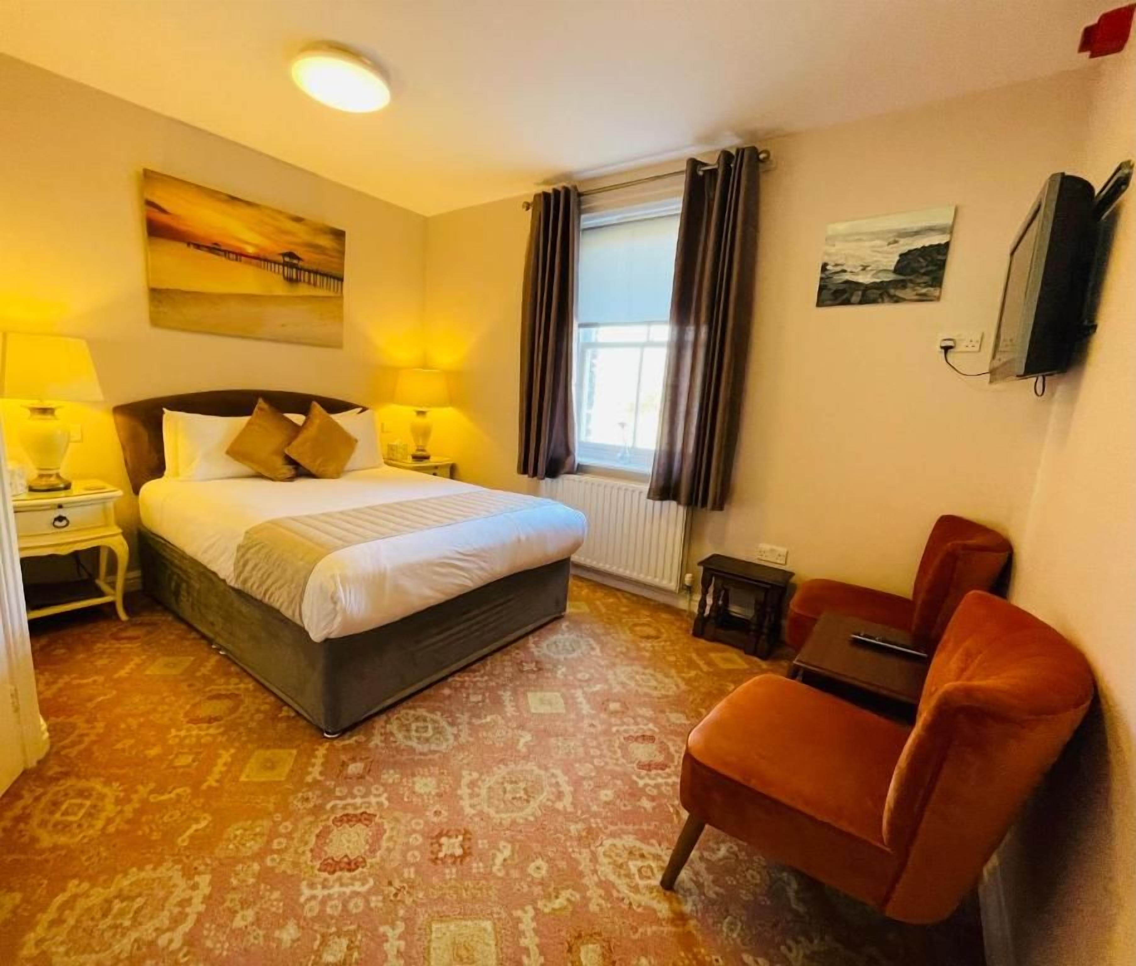 A hotel room with a bed, two bedside lamps, a chair, a small table, a window with curtains, and artwork on the walls.