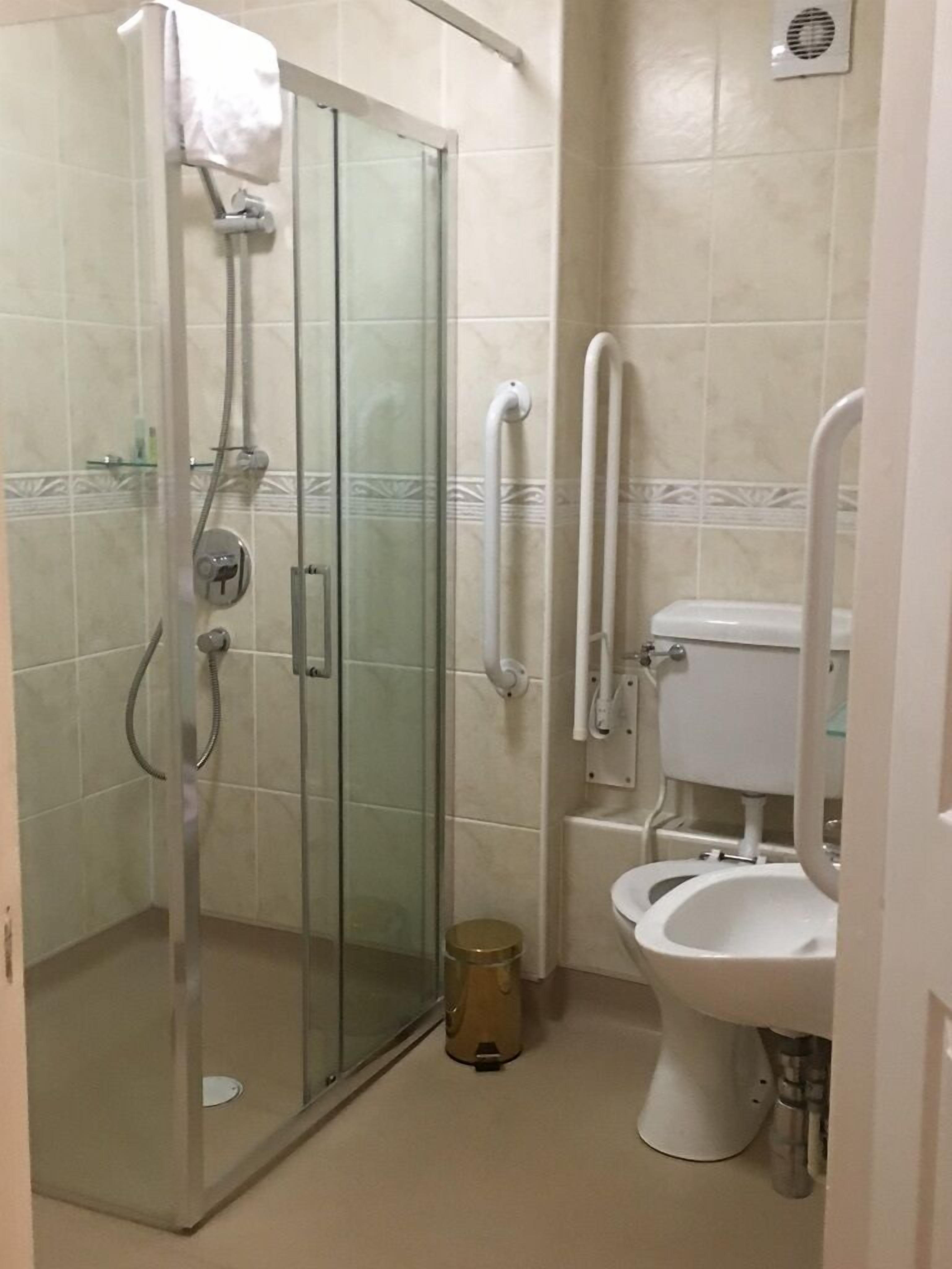 Un baño con ducha de vidrio, inodoro con asiento elevado y barra de apoyo.