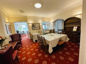 Un coin repas avec des tables disposées pour accueillir quatre personnes, une moquette rouge à motifs et un buffet en bois contenant des verres.