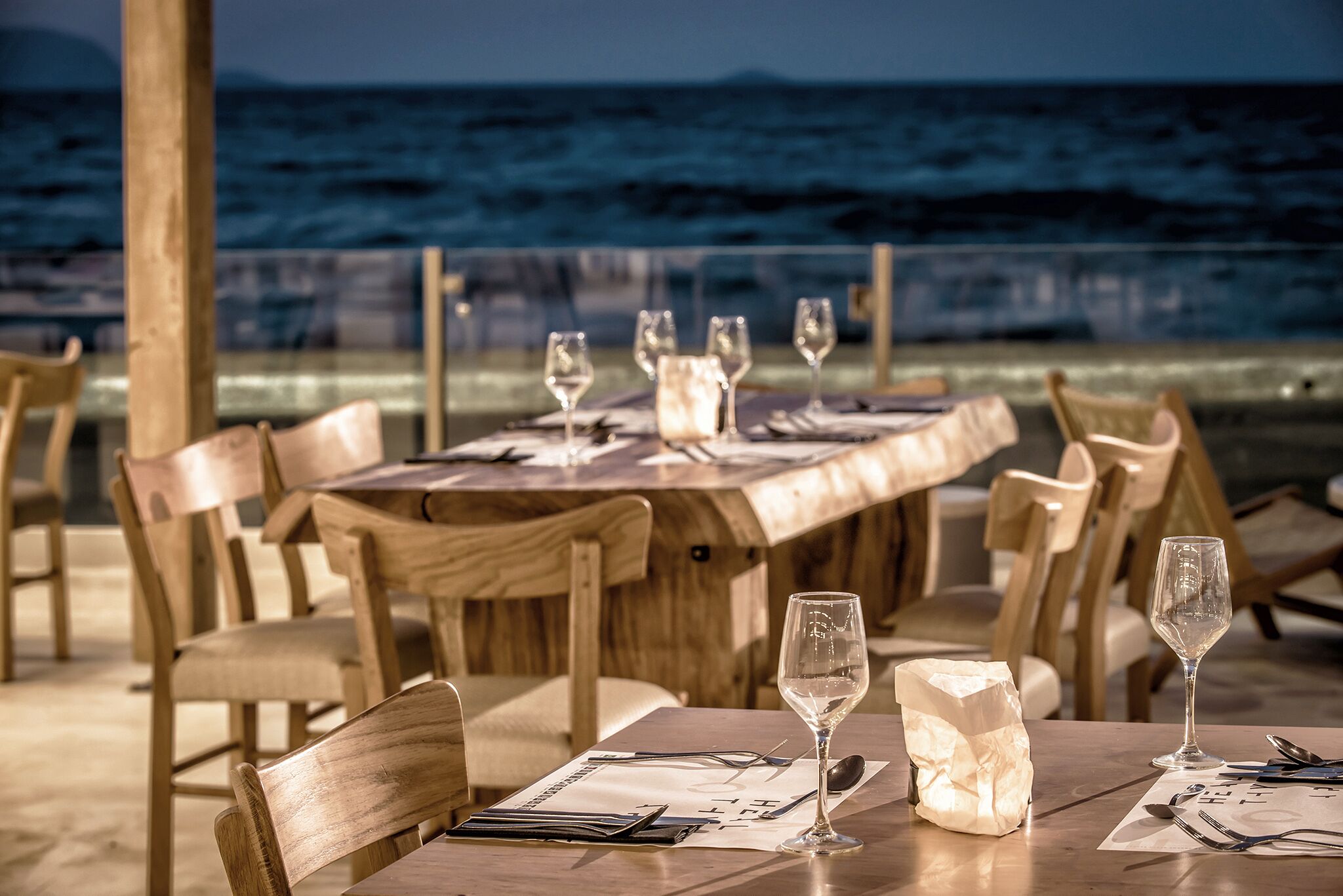 A wooden dining table set with wine glasses and cutlery, overlooking the ocean.