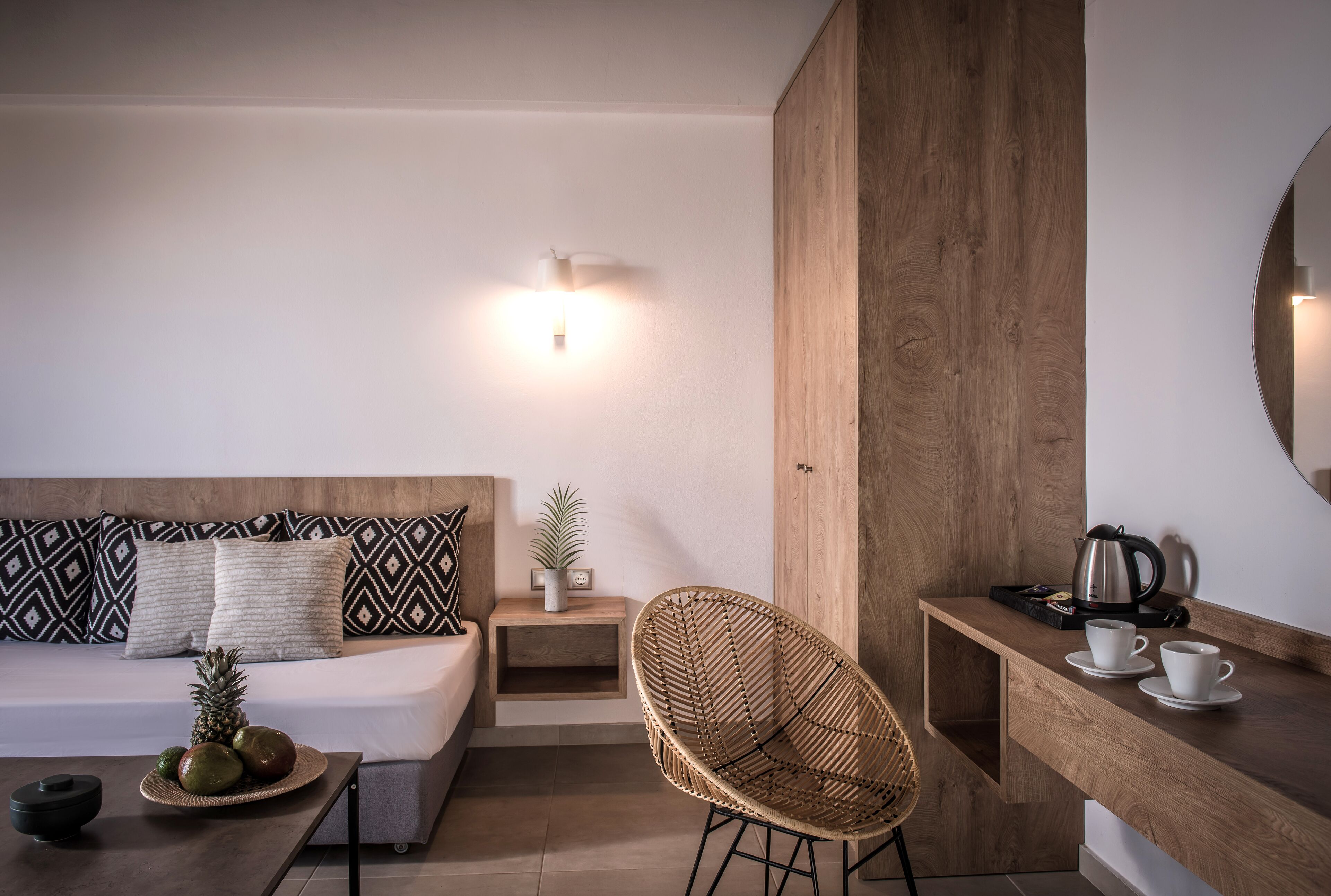 Een moderne hotelkamer met een houten hoofdbord, een ronde spiegel, een rieten stoel, een klein nachtkastje met een plant, en een dienblad met kopjes en een waterkoker.