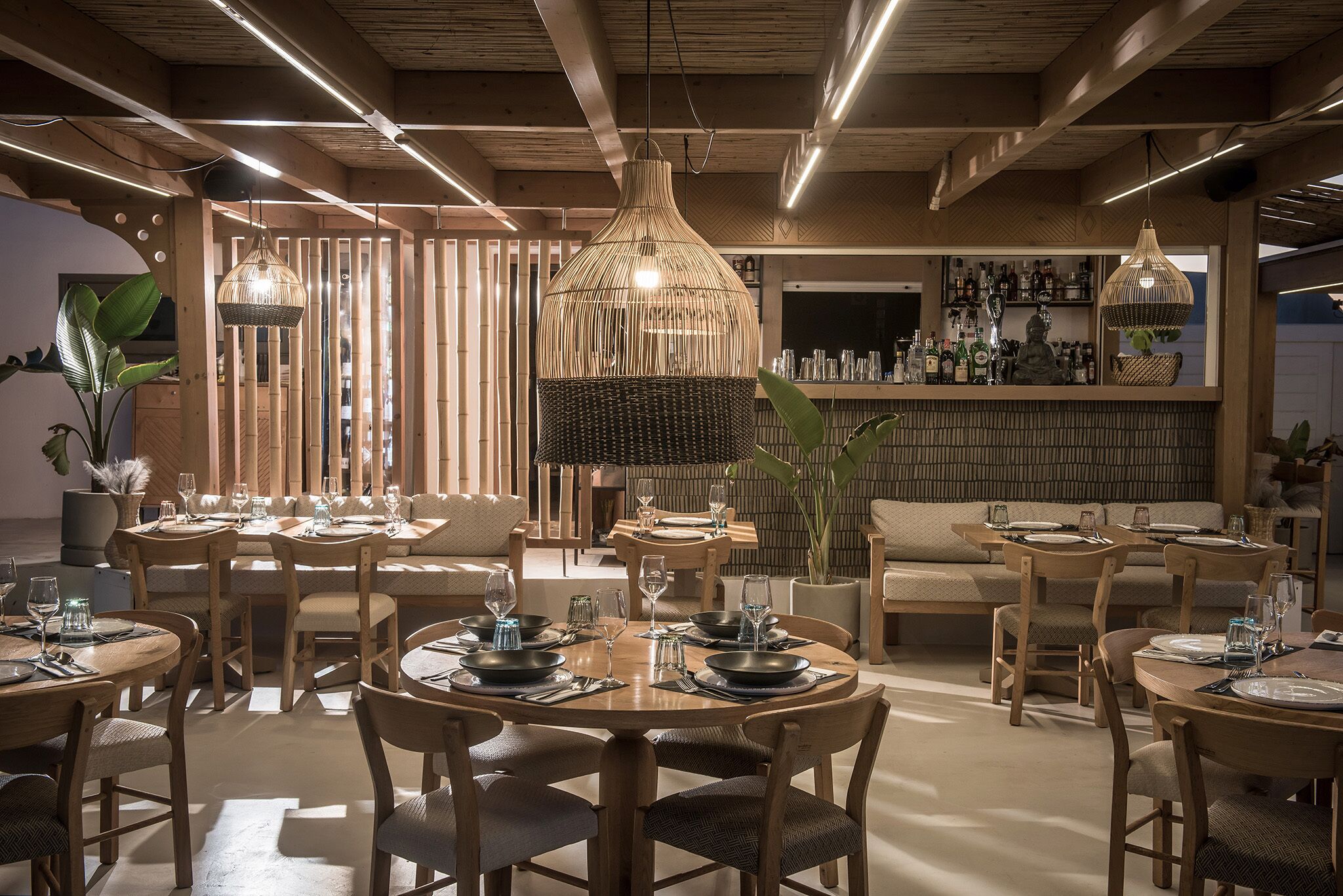 A modern dining area with round tables, wicker pendant lights, and a bar in the background.