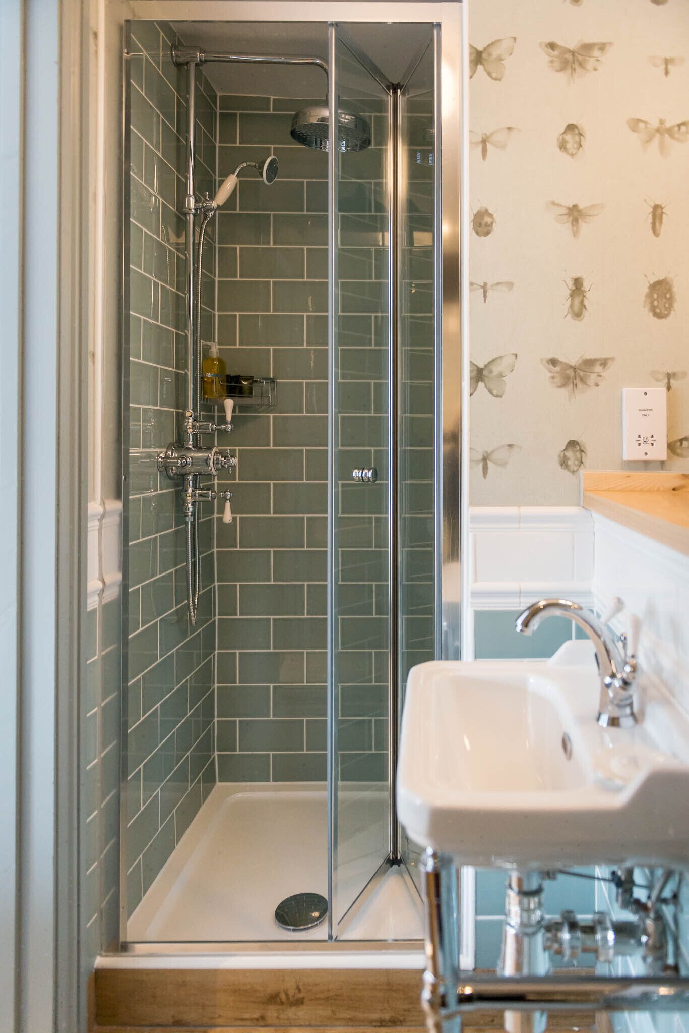 A glass-enclosed shower with a tiled wall, a sink, and a butterfly-themed wallpaper.