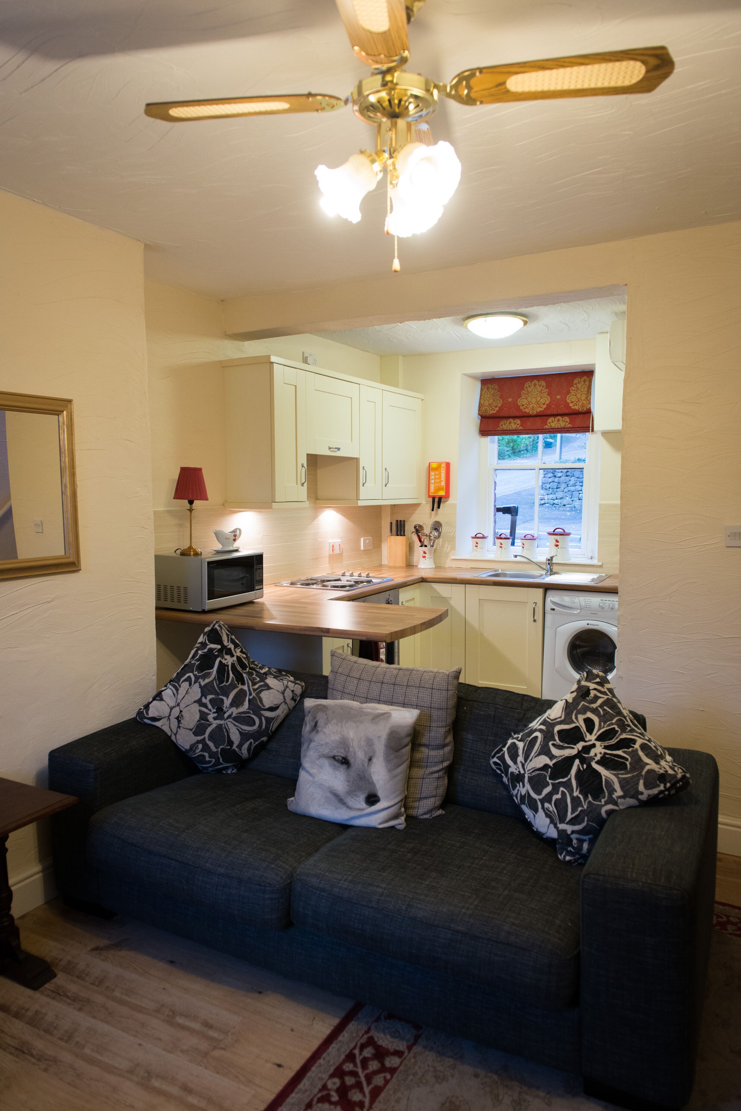 A compact living space with a sofa, a small table, a ceiling fan, and a kitchen area with cabinets and a washing machine.