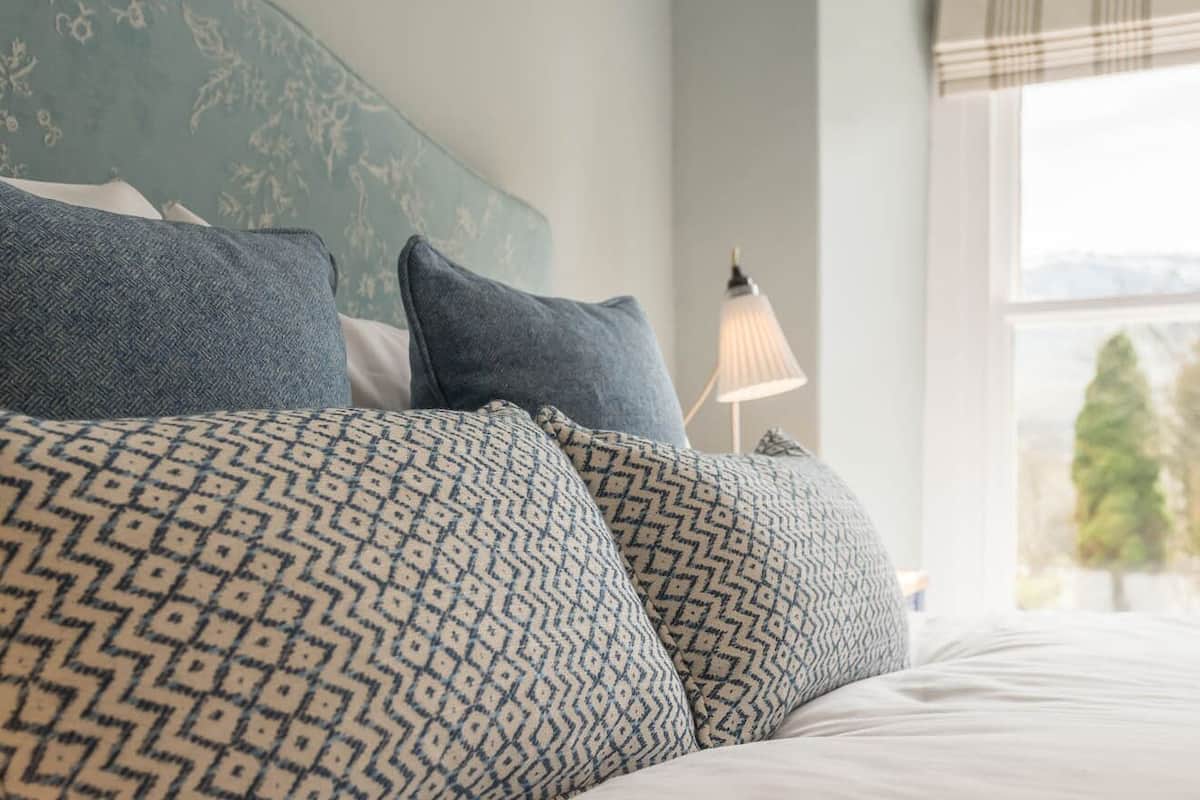 A bed with patterned pillows, a lamp, and a window with a view of greenery.