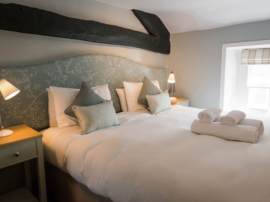 Una cama bien hecha con sábanas blancas y varias almohadas, una lámpara de noche y una ventana con persianas.