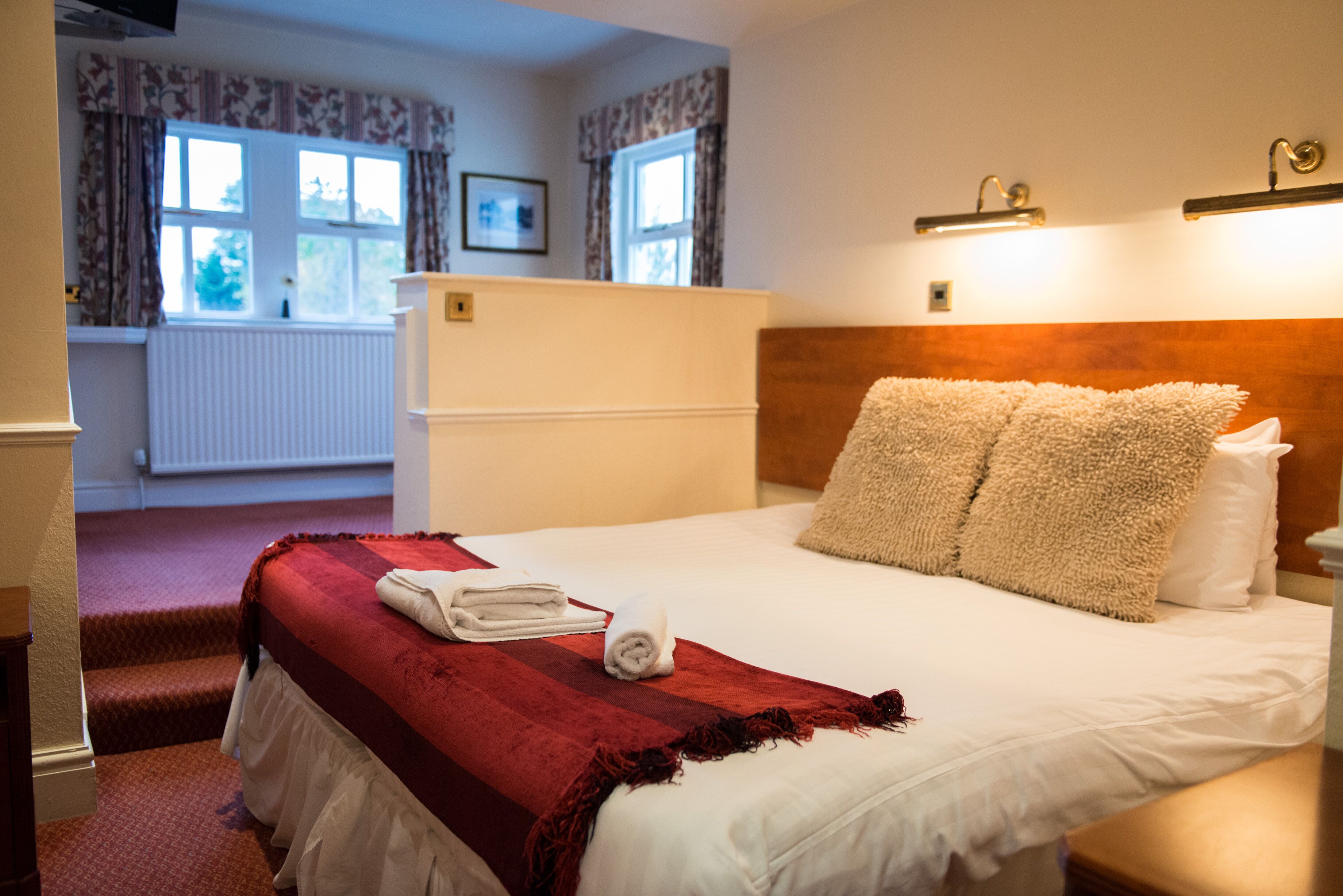 Une chambre d’hôtel avec un lit, un plaid rouge, des oreillers blancs, une tête de lit en bois et une fenêtre avec des rideaux.