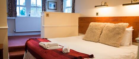 A hotel room with a bed, a red throw blanket, white pillows, a wooden headboard, and a window with curtains.