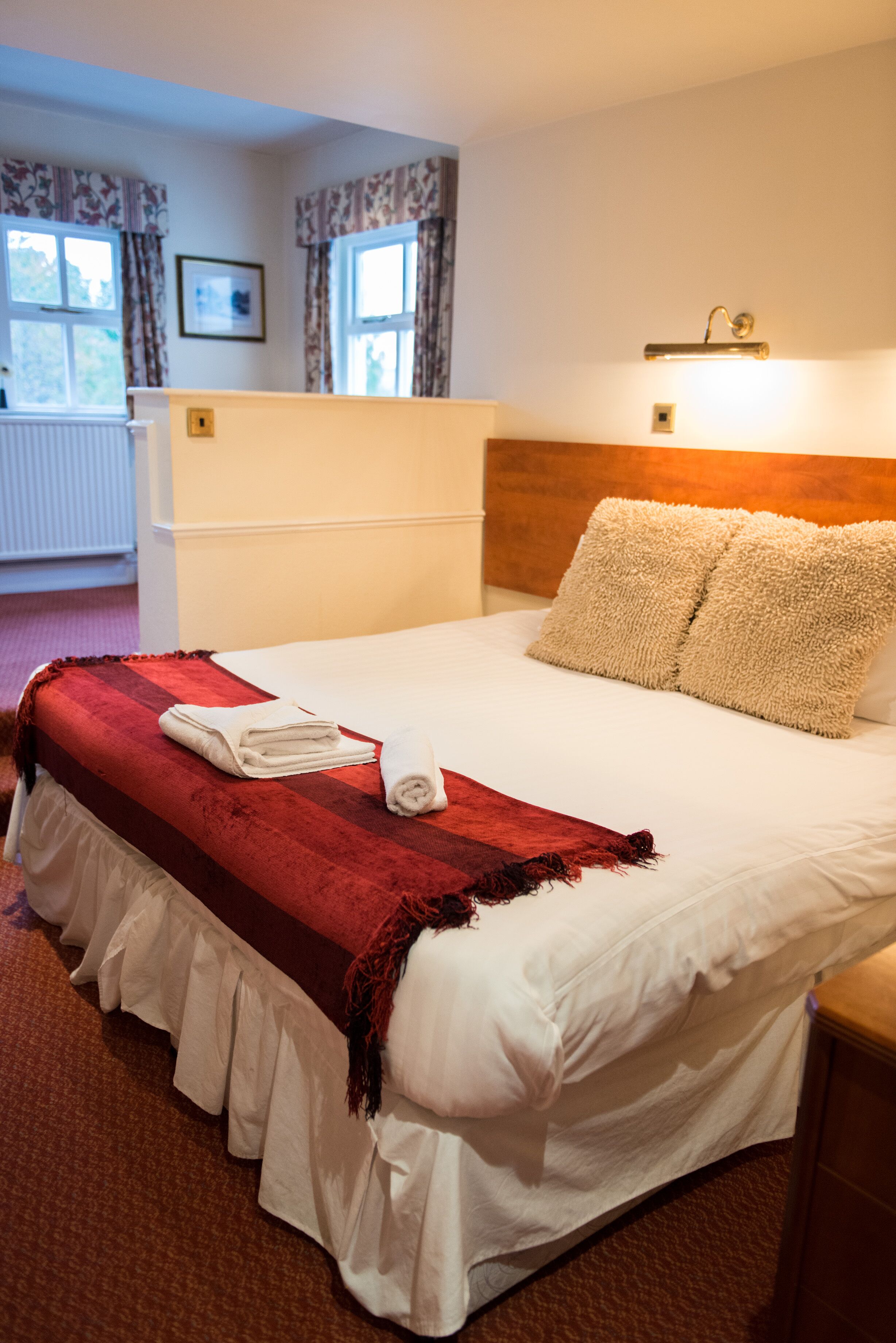 A neatly made bed with a red blanket, white pillows, and a white duvet. There is a wooden headboard, a bedside table, and a lamp on the wall.
