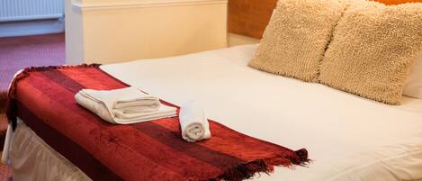 A neatly made bed with a red blanket, white pillows, and a white duvet. There is a wooden headboard, a bedside table, and a lamp on the wall.