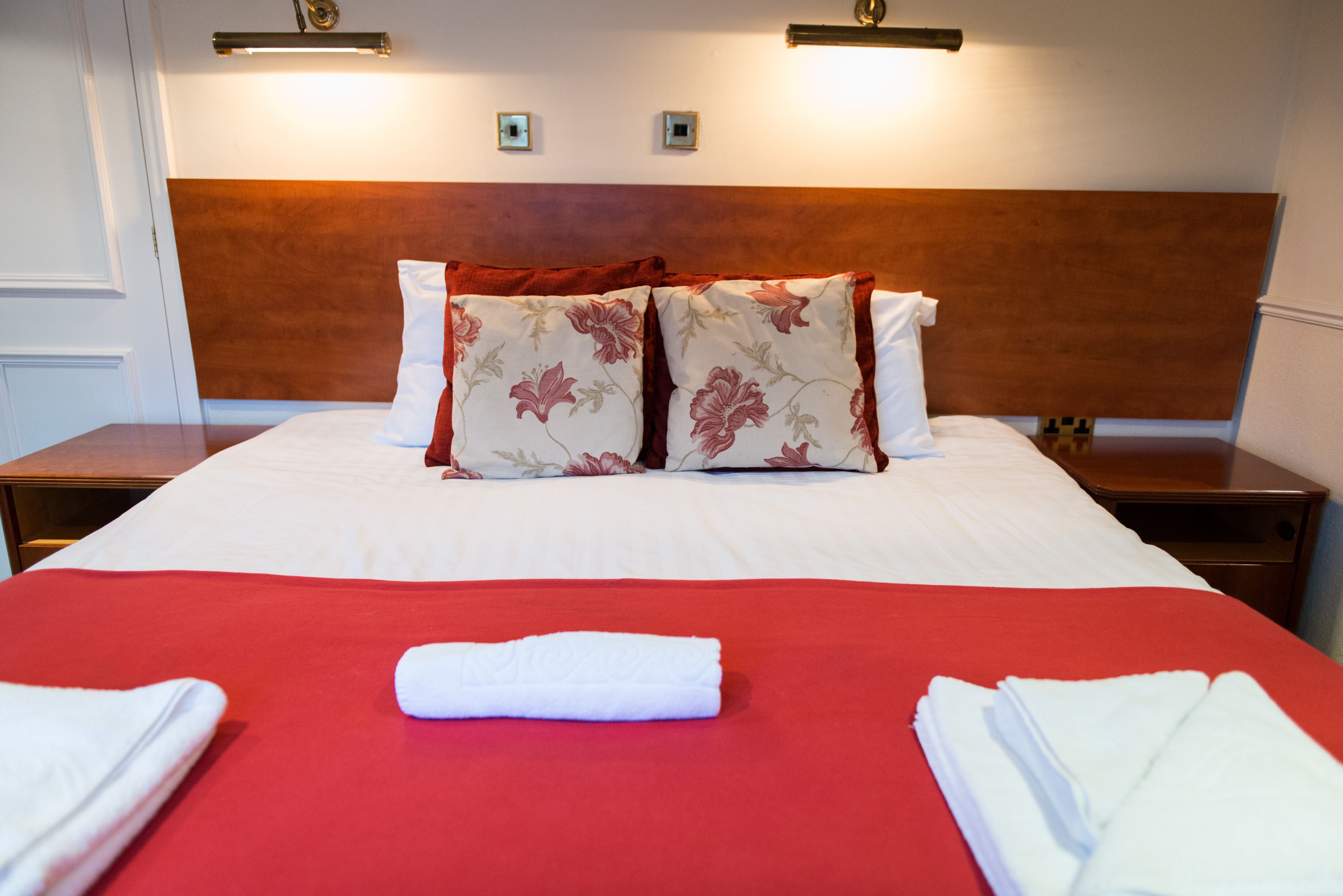 Un letto rifatto con una coperta rossa, due cuscini floreali e un asciugamano bianco piegato.