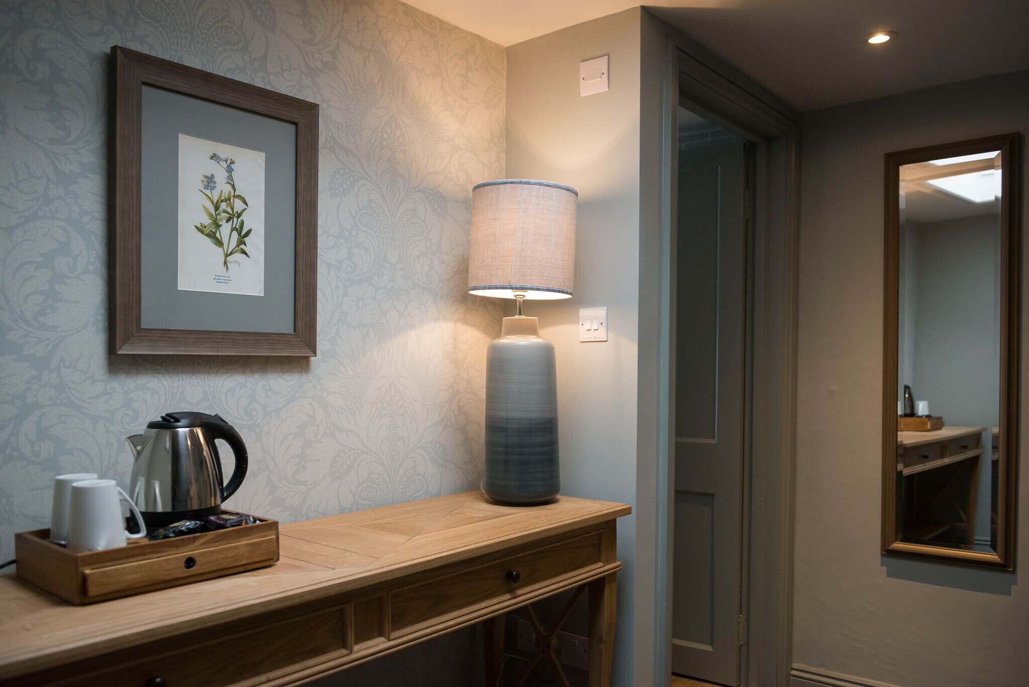 A wooden console table with a framed botanical print, a lamp, and a kettle.
