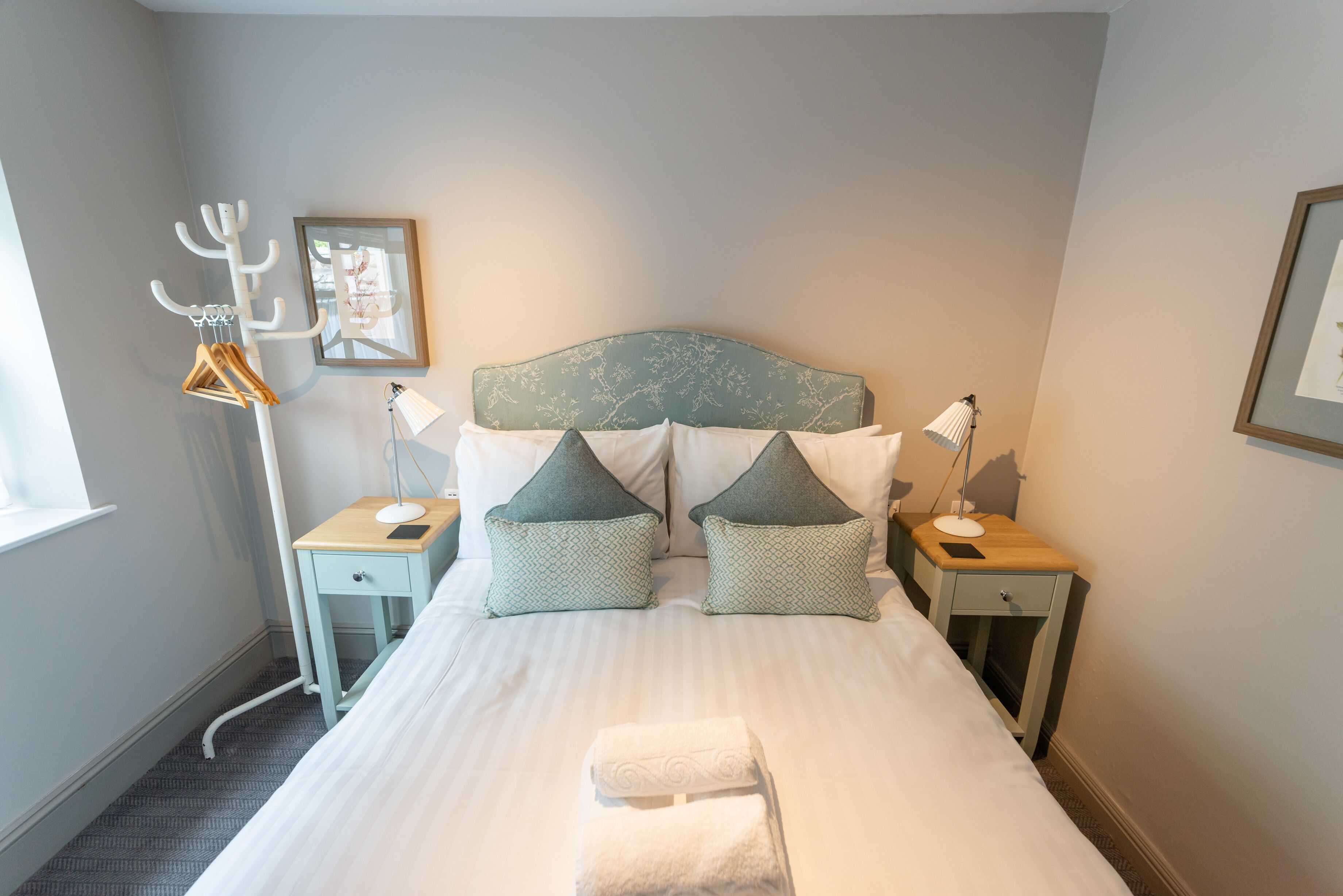A neatly made bed with a headboard, two bedside tables with lamps, a coat rack, and a framed picture on the wall.