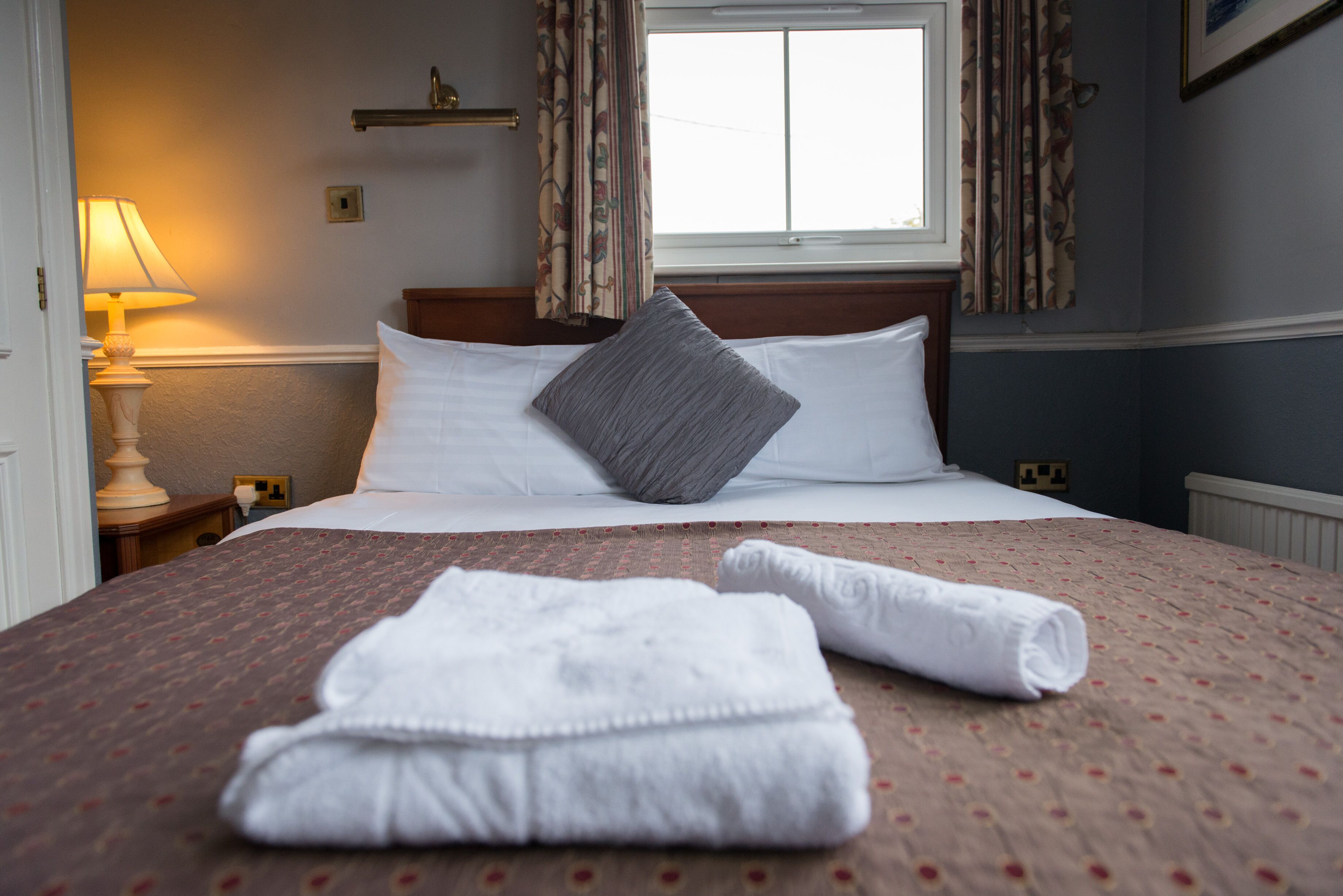 A neatly made bed with white linens and a grey pillow, a rolled white towel on the bed, and a lamp on a bedside table.