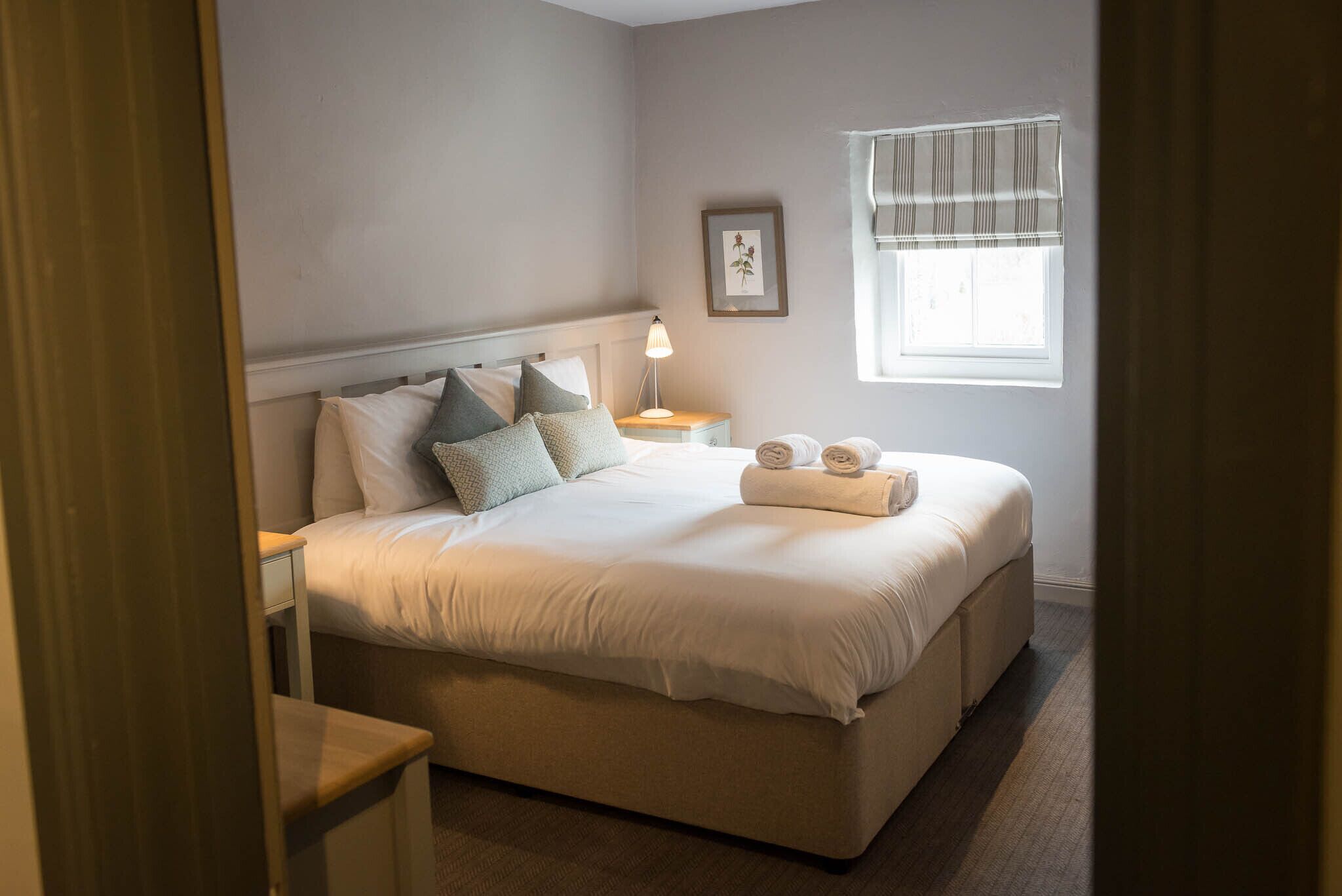 Una cama bien hecha con almohadas y una toalla doblada, una mesita de noche con una lámpara, un cuadro en la pared y una ventana con persianas.
