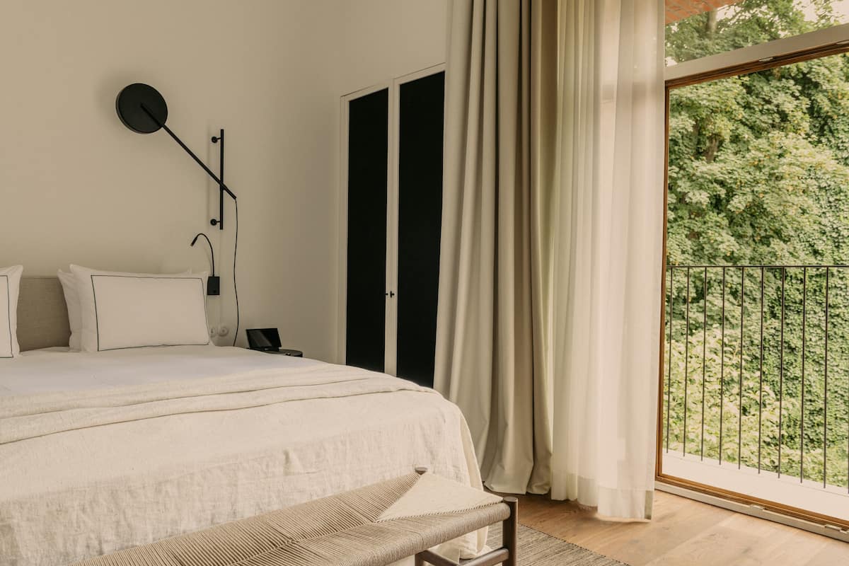 Un dormitorio con una cama grande, una mesita de noche, una lámpara y vistas a un balcón con vegetación.