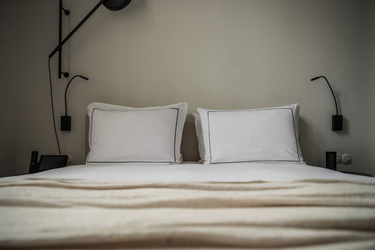 Una cama bien hecha con ropa de cama blanca y dos almohadas, una mesita de noche con una lámpara y un enchufe de pared.