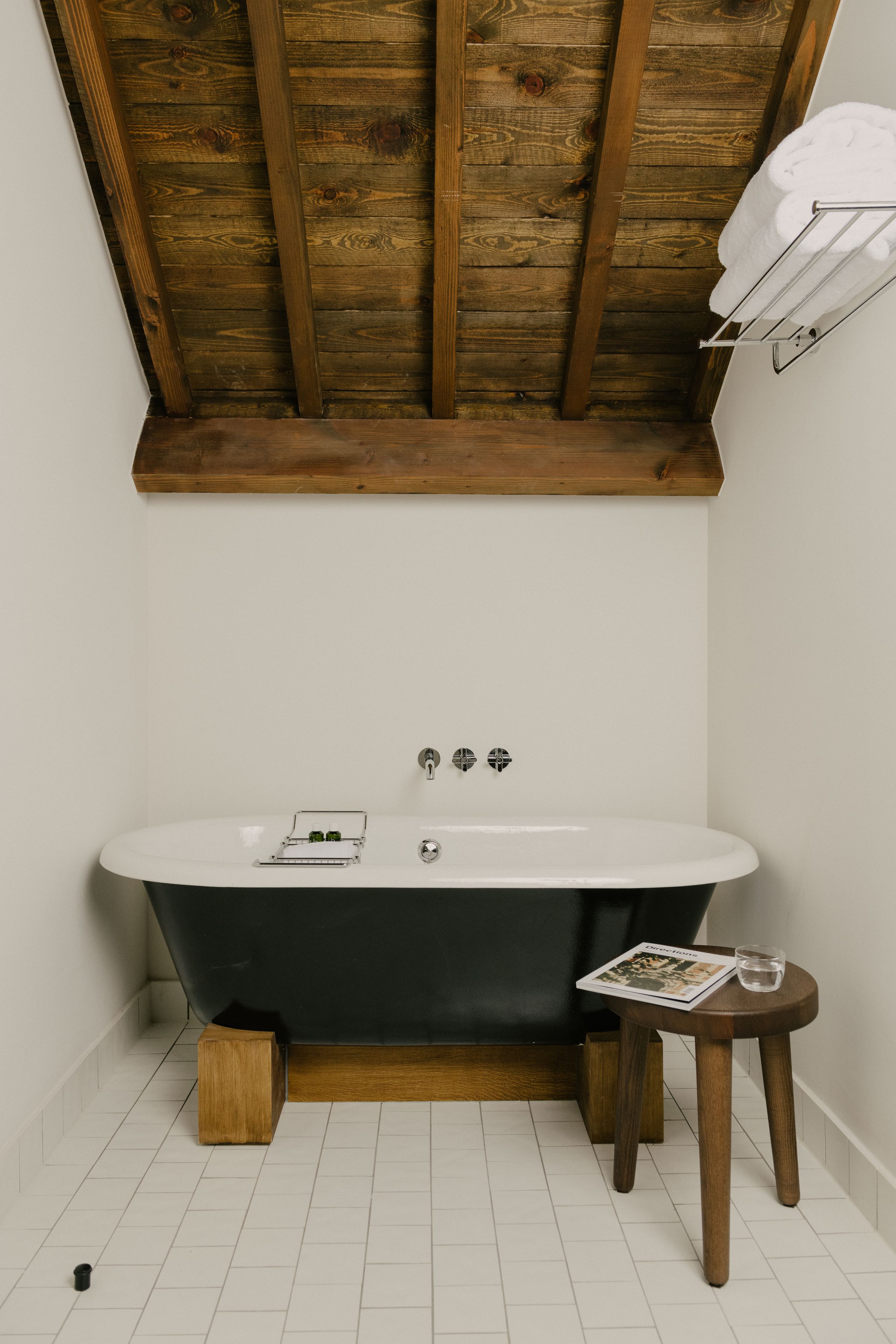 A bathroom with a freestanding bathtub, wooden beams on the ceiling, and a small wooden stool with a magazine and glass of water.