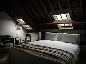 A bedroom with a sloped ceiling, skylights, a bed, a bedside table, a chair, and a lamp.