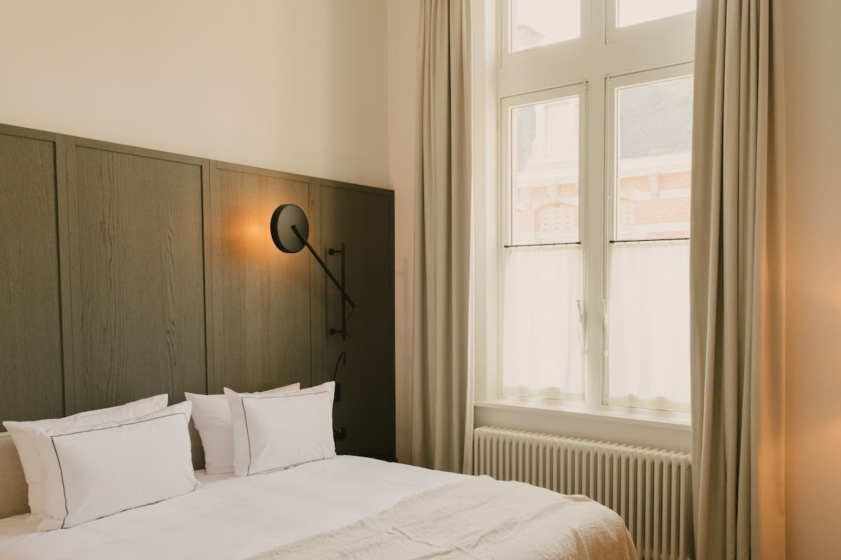 Habitación de hotel con cabecera de madera, cama con ropa blanca, lámpara de pared y ventana con cortinas.