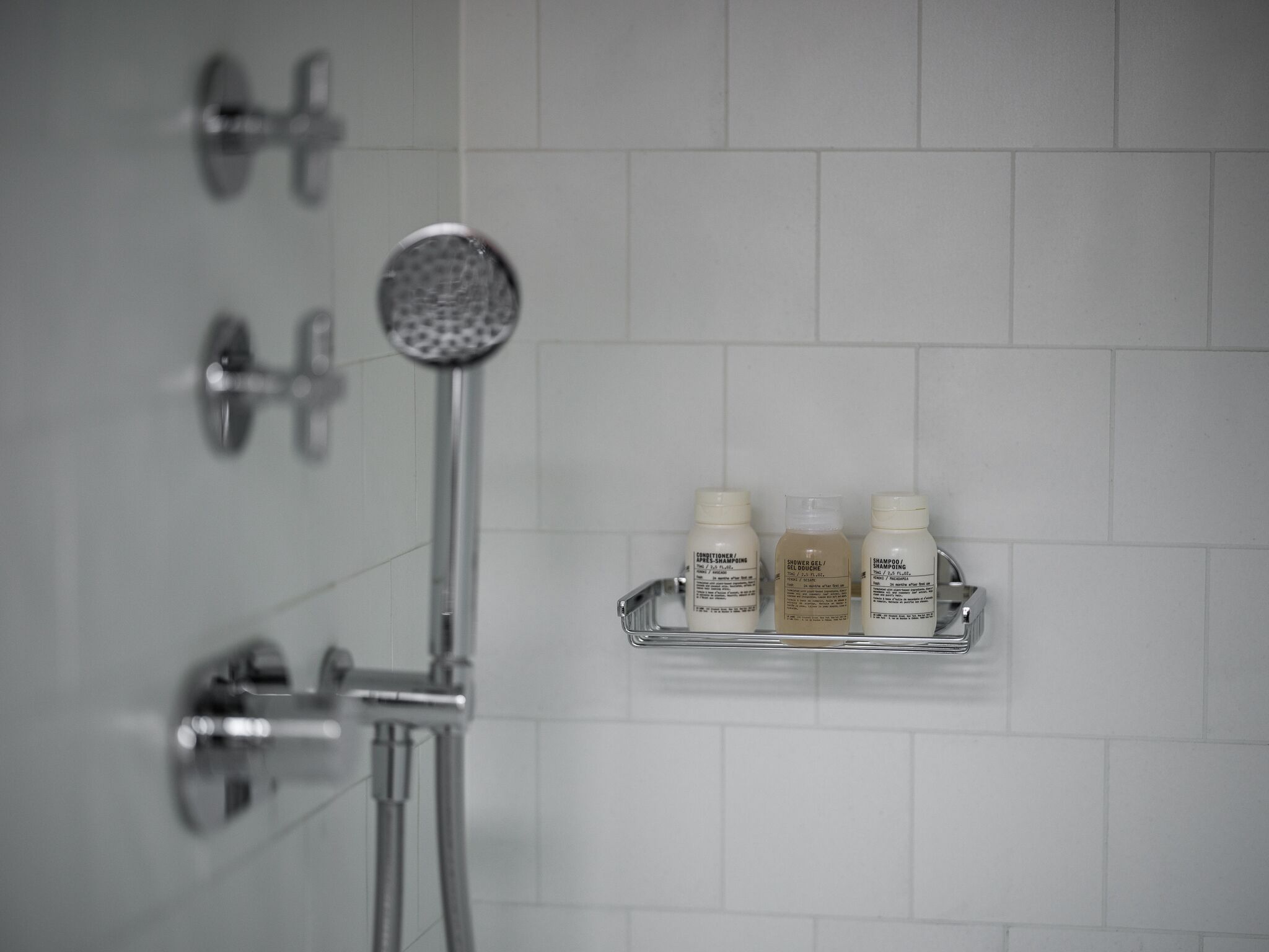 A shower with a handheld showerhead and a wall-mounted faucet. There are three bottles of toiletries on a shelf: conditioner, shower gel, and shampoo.