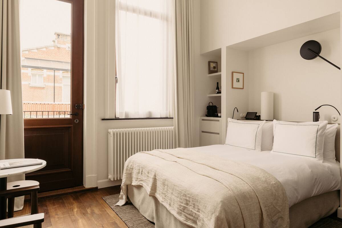 Habitación de hotel con cama, escritorio, silla, radiador, ventana con cortinas y vistas a edificios.