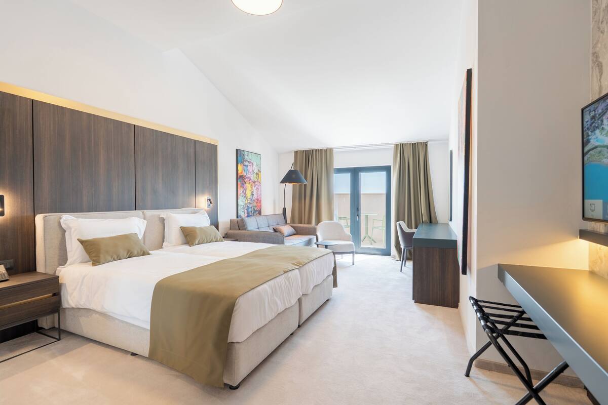 A modern hotel room with a large bed, a desk, a chair, and a balcony with curtains.