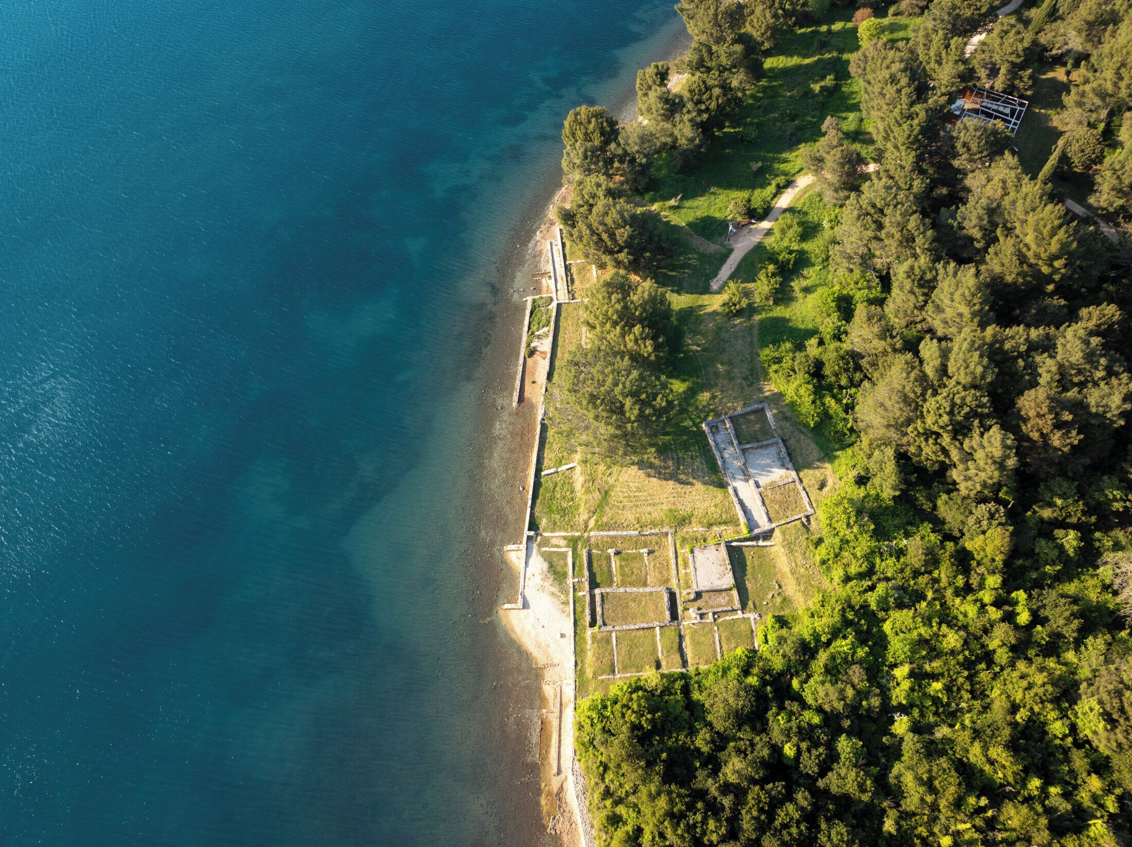 An aerial view of a coastal property with a beach, dense forest, and a large rectangular building.
