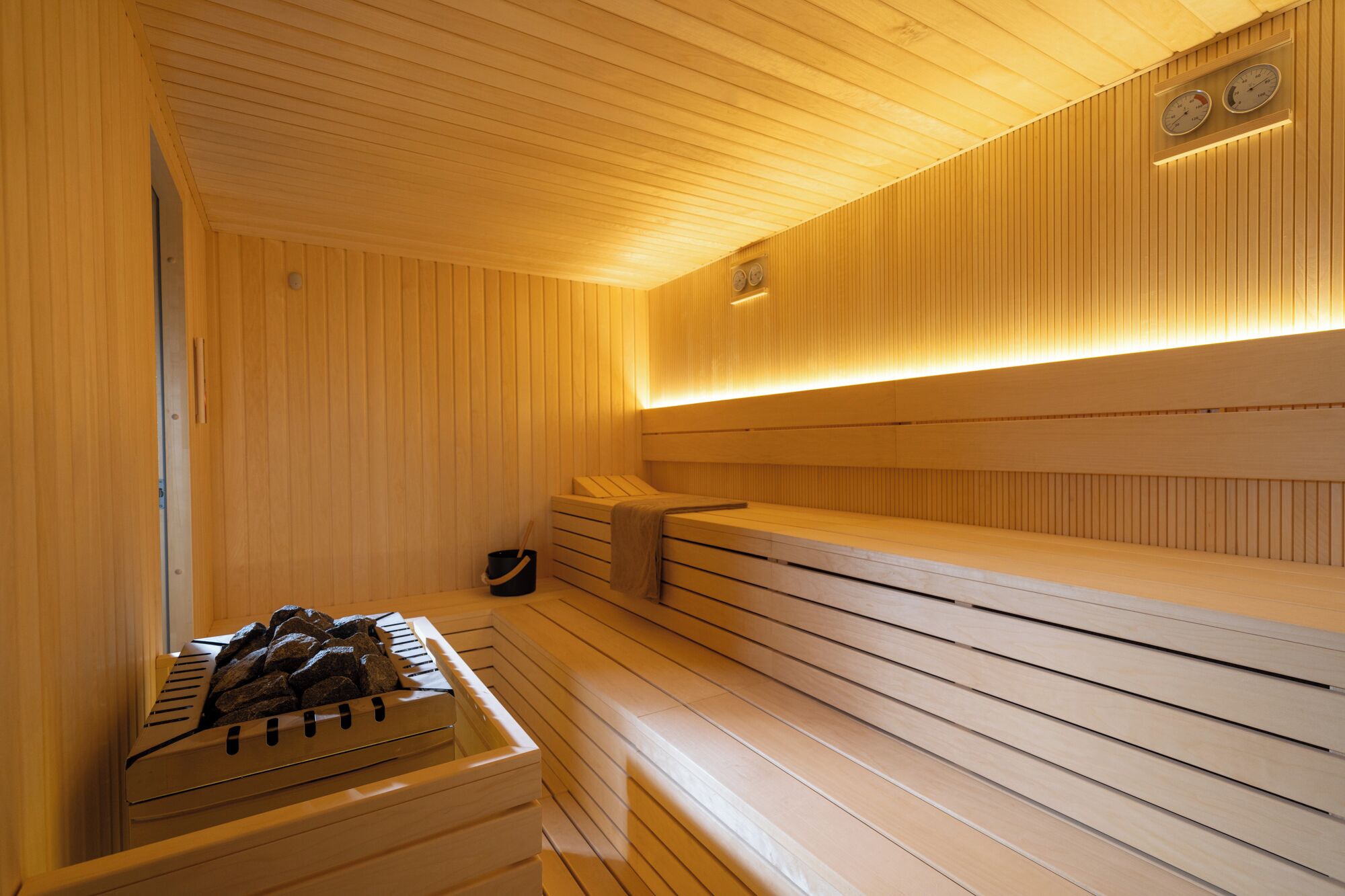 Un sauna avec des bancs en bois, un banc muni d’une serviette et un récipient rempli de pierres.