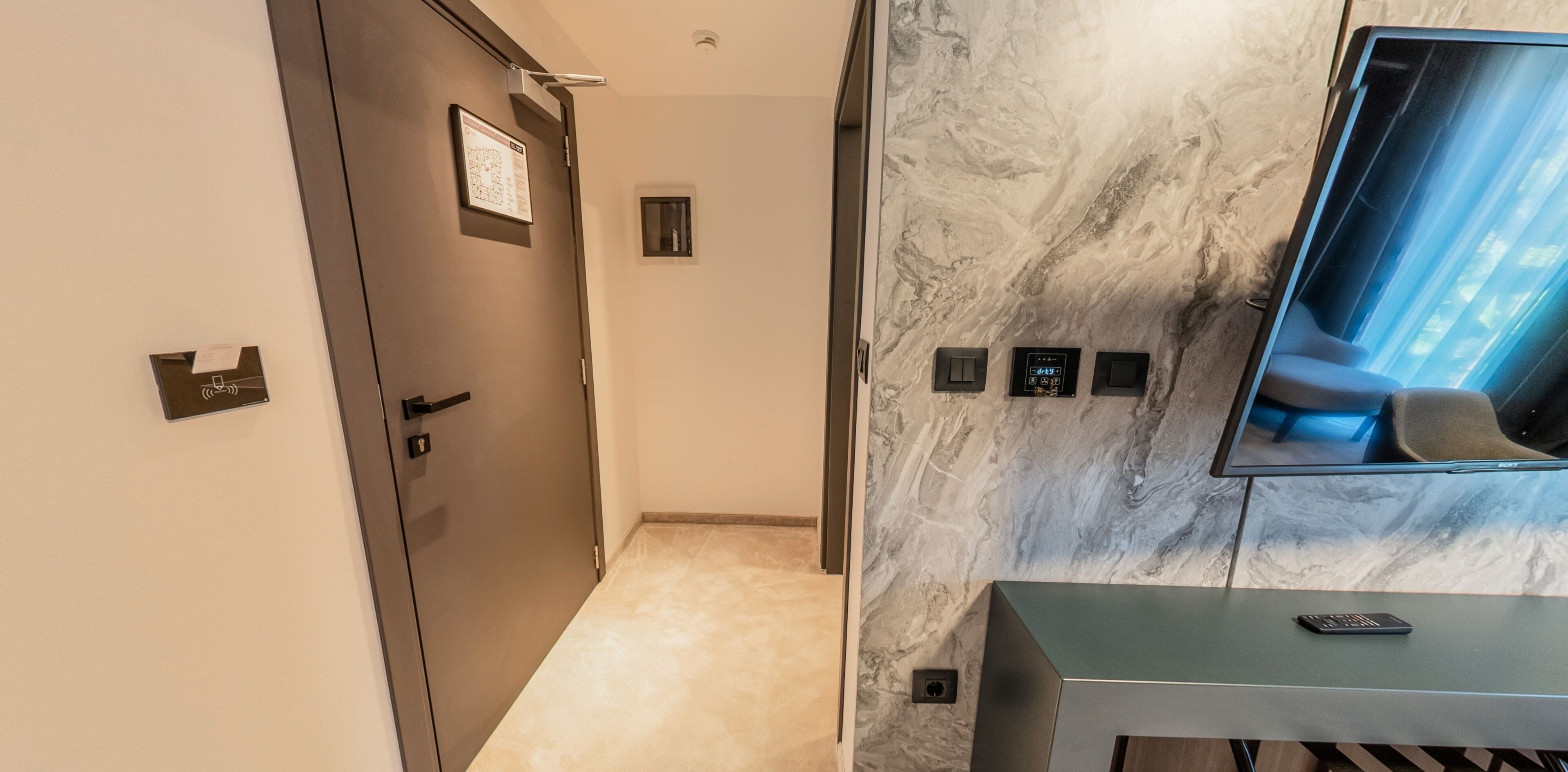 Een moderne hotelgang met een marmeren wand, een deur, een klein raam en een bedieningspaneel.
