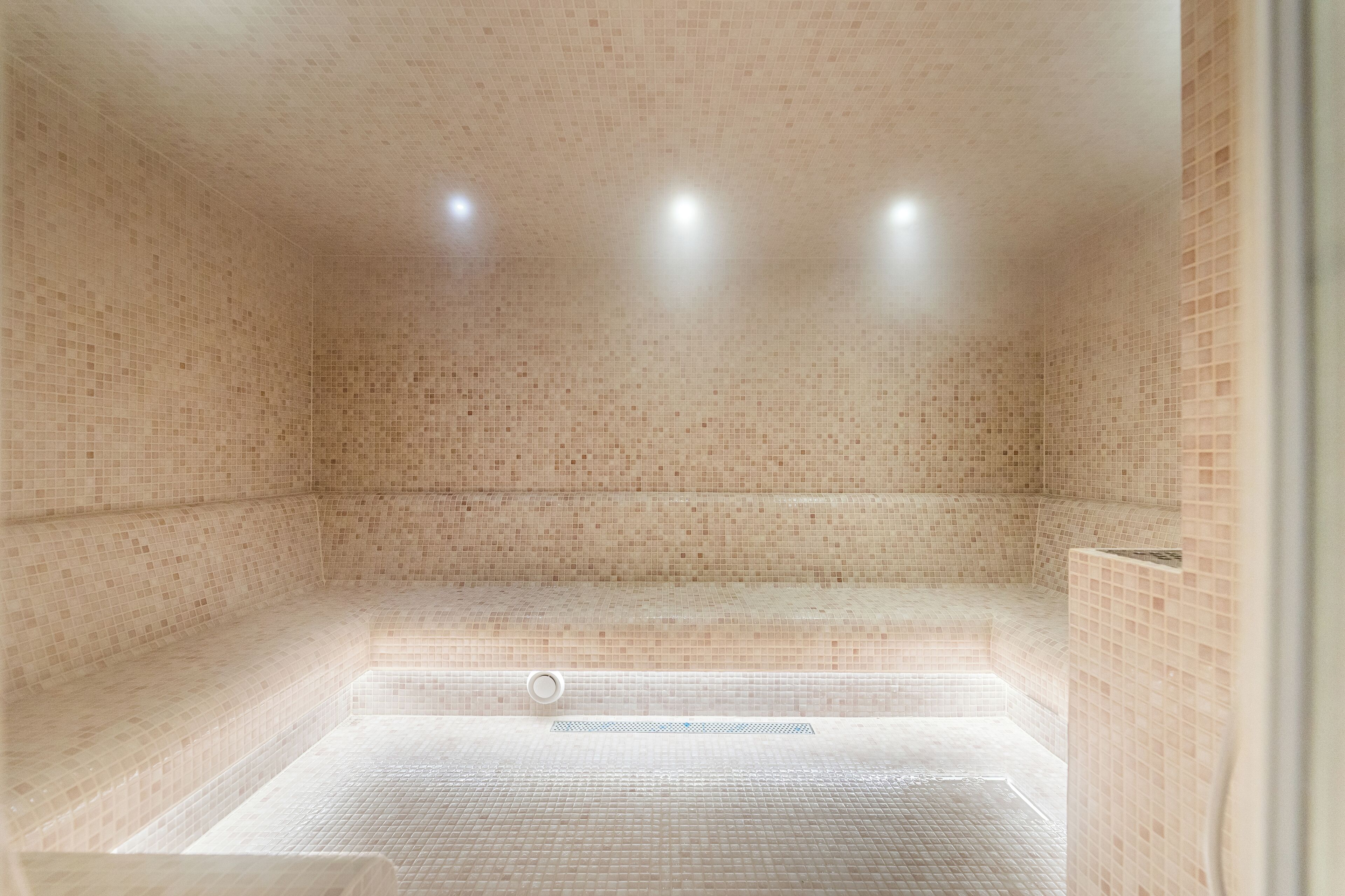 A tiled room with a bench, a drain, and three ceiling lights.