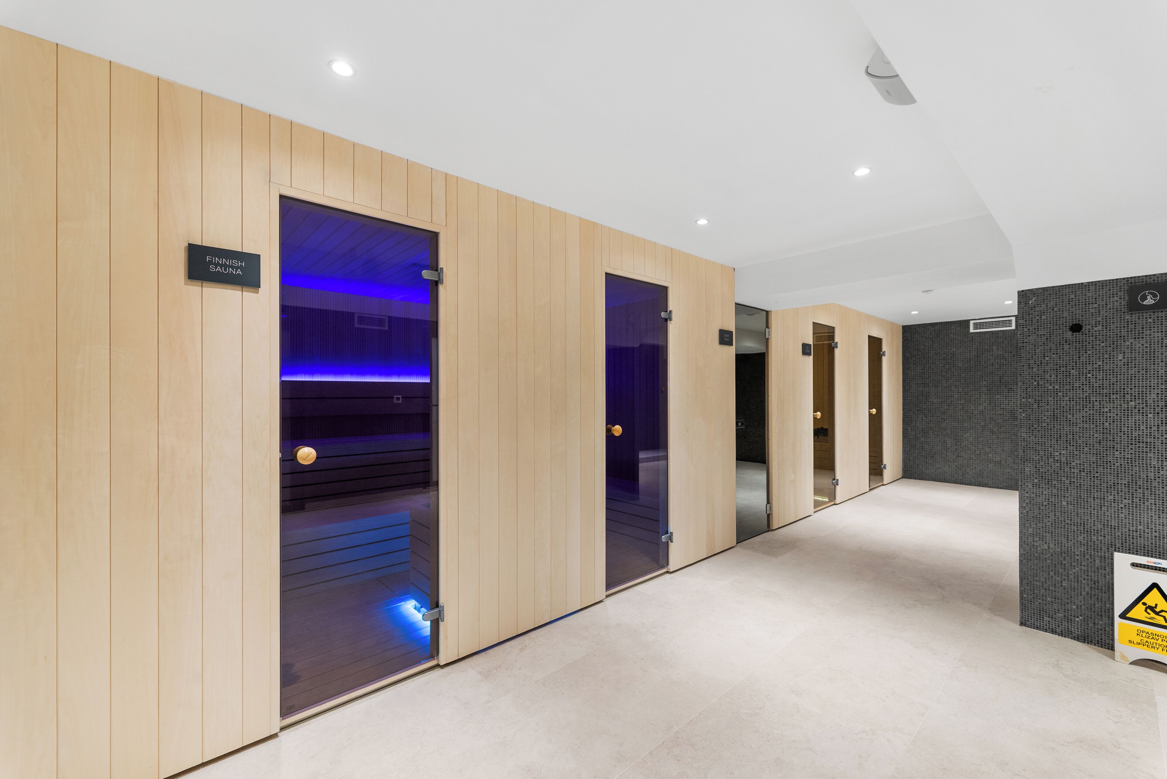 Un couloir aux murs recouverts de lambris en bois, un panneau indiquant la présence d’un sauna finlandais et un panneau d’avertissement au sol.