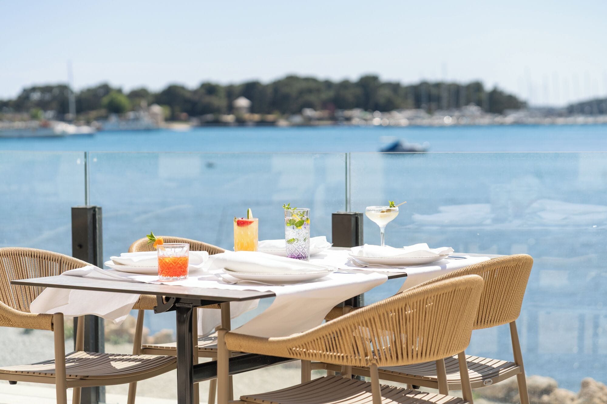 A dining table with chairs, a glass of orange juice, a tall glass with mint, and a martini glass, set against a view of a calm body of water and a distant shoreline.
