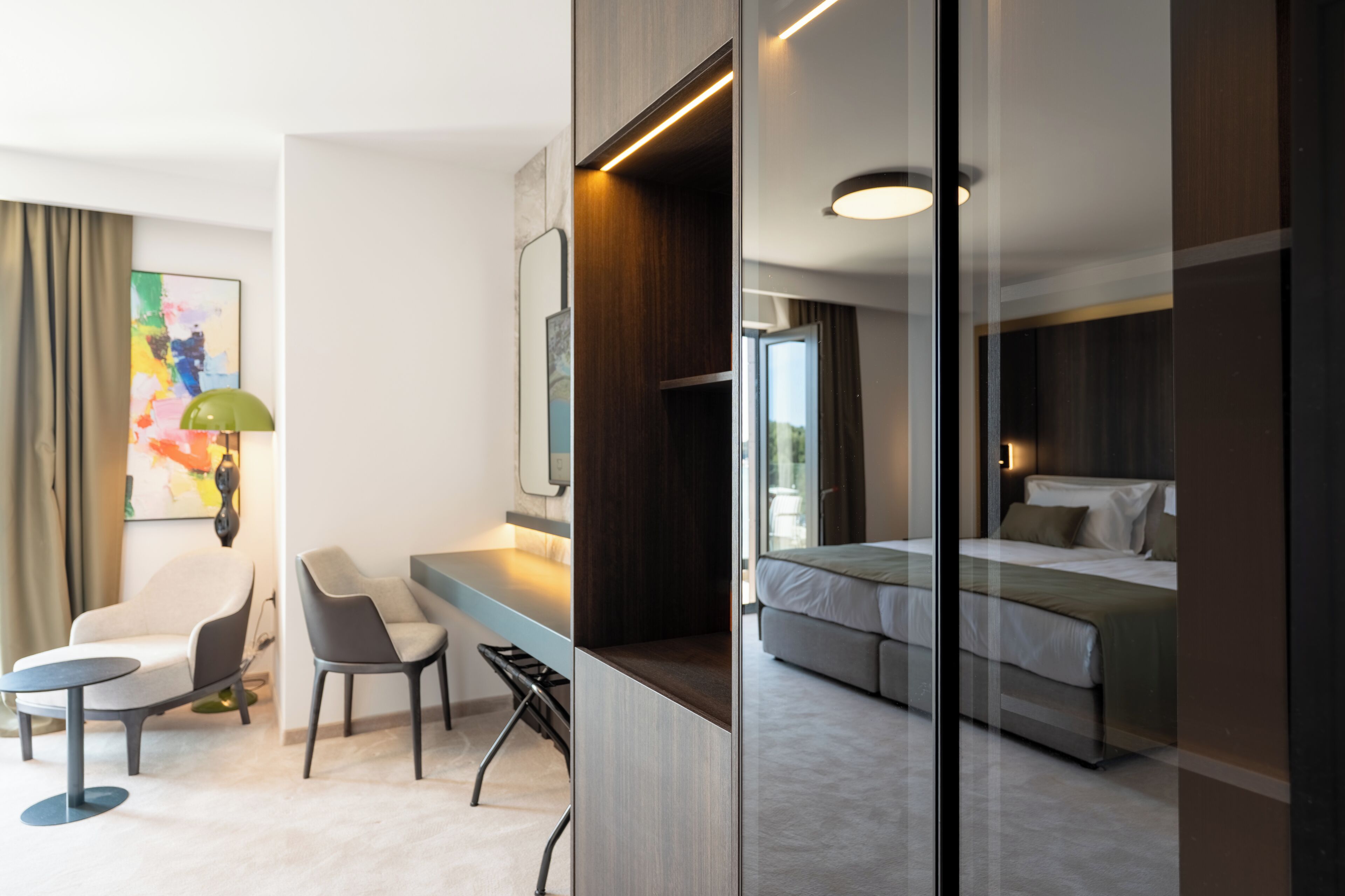 A modern hotel room with a bed, a desk, two chairs, a desk lamp, and a large abstract painting on the wall.