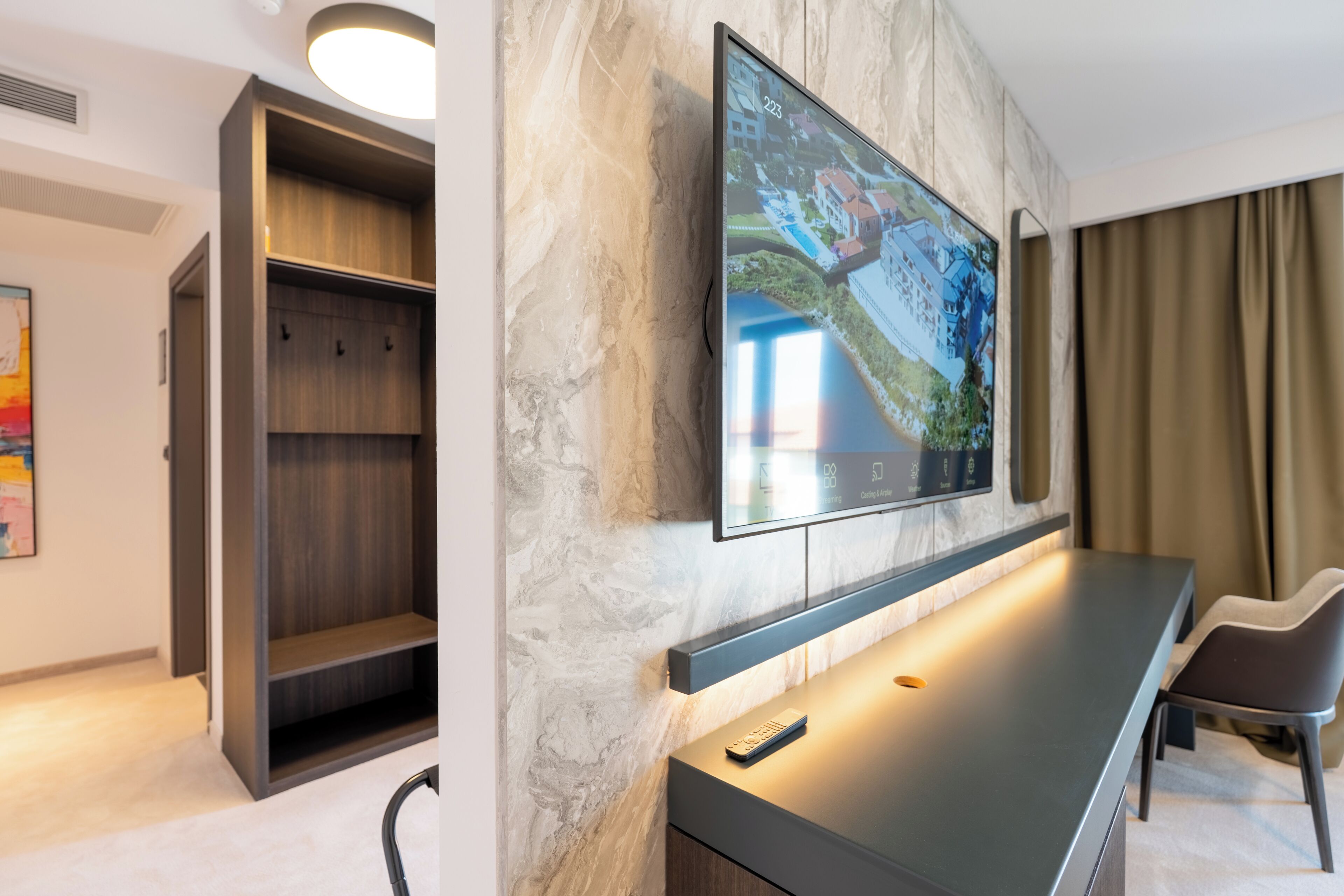 Ett modernt hotellrum med en stor platt-TV som  är monterad på en vägg av marmor, en inbyggd bänk och en garderob i trä.