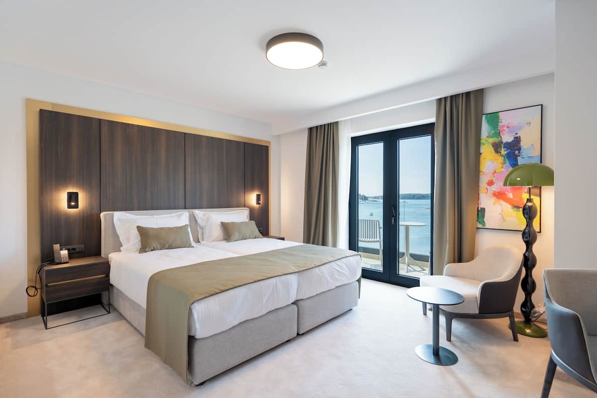 A modern hotel room with a large bed, bedside tables, a chair, a small table, and a view of the sea.