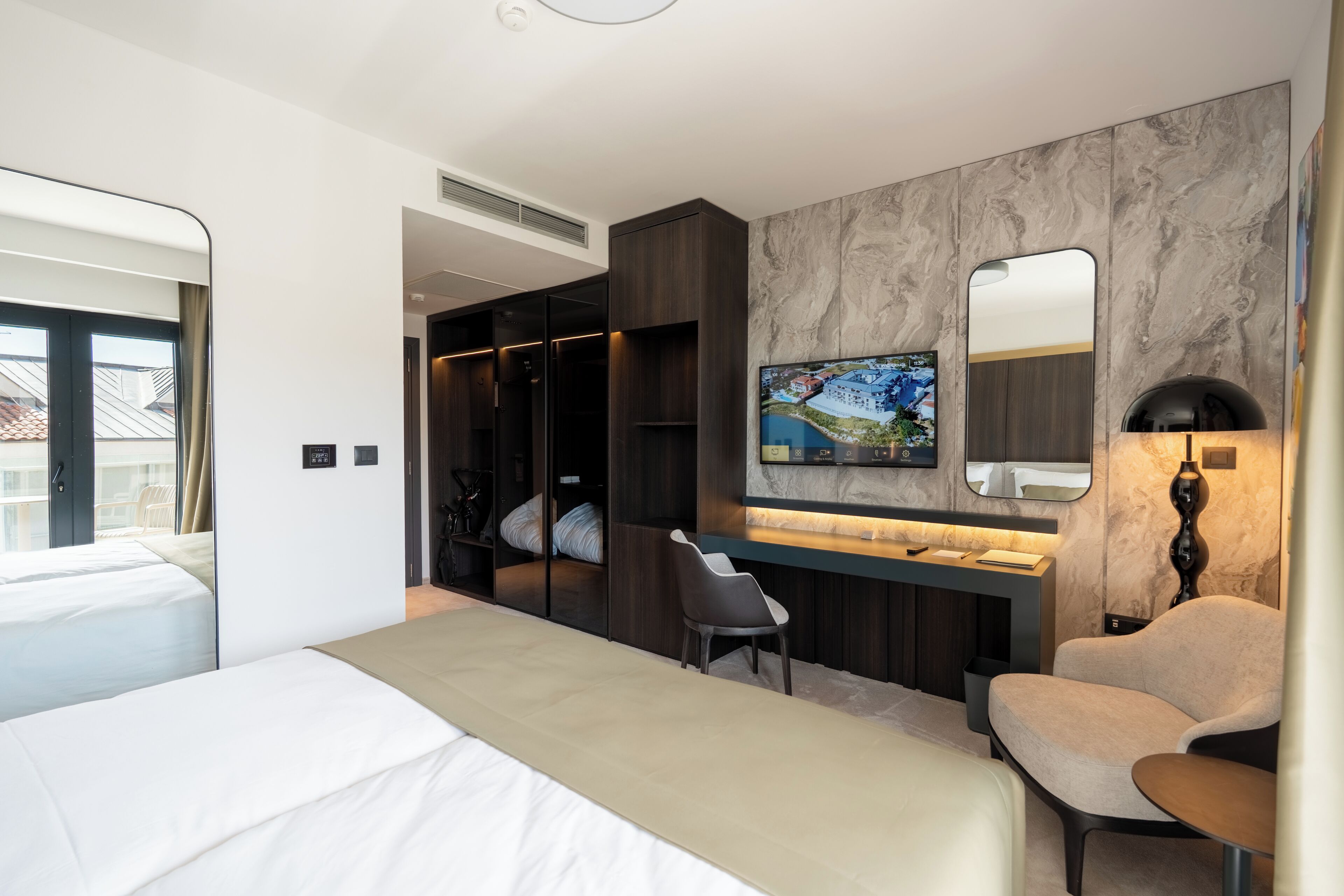 A modern hotel room with a bed, a desk, a chair, a TV, and a wardrobe.