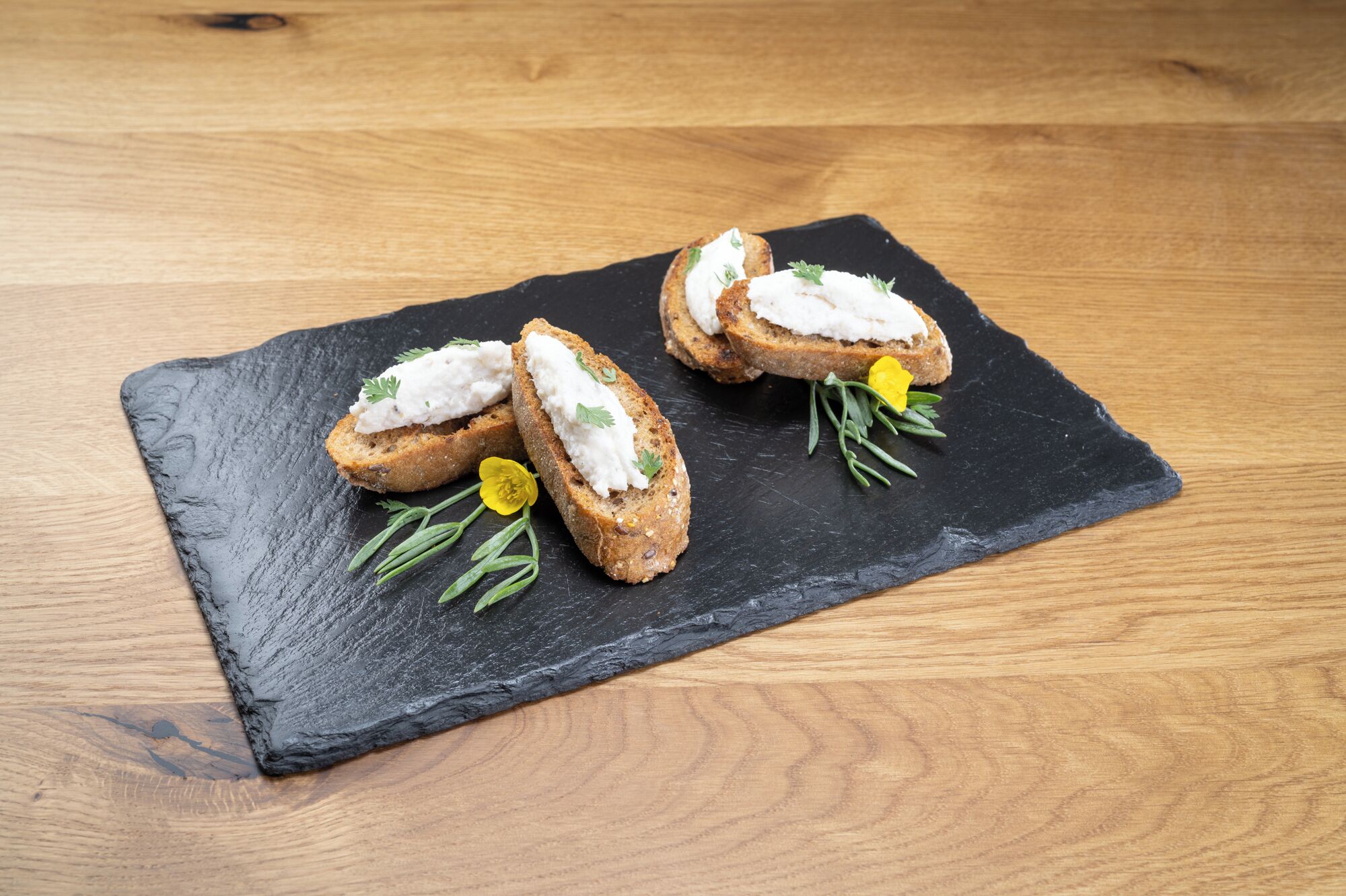 A black slate serving board with three pieces of bread topped with a white spread and garnished with herbs, placed on a wooden table.