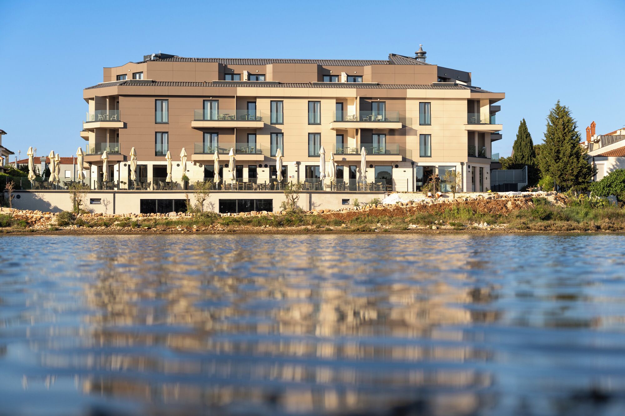 Een meerlaags hotel met balkons, buiten zitplaatsen en uitzicht op een wateroppervlak.