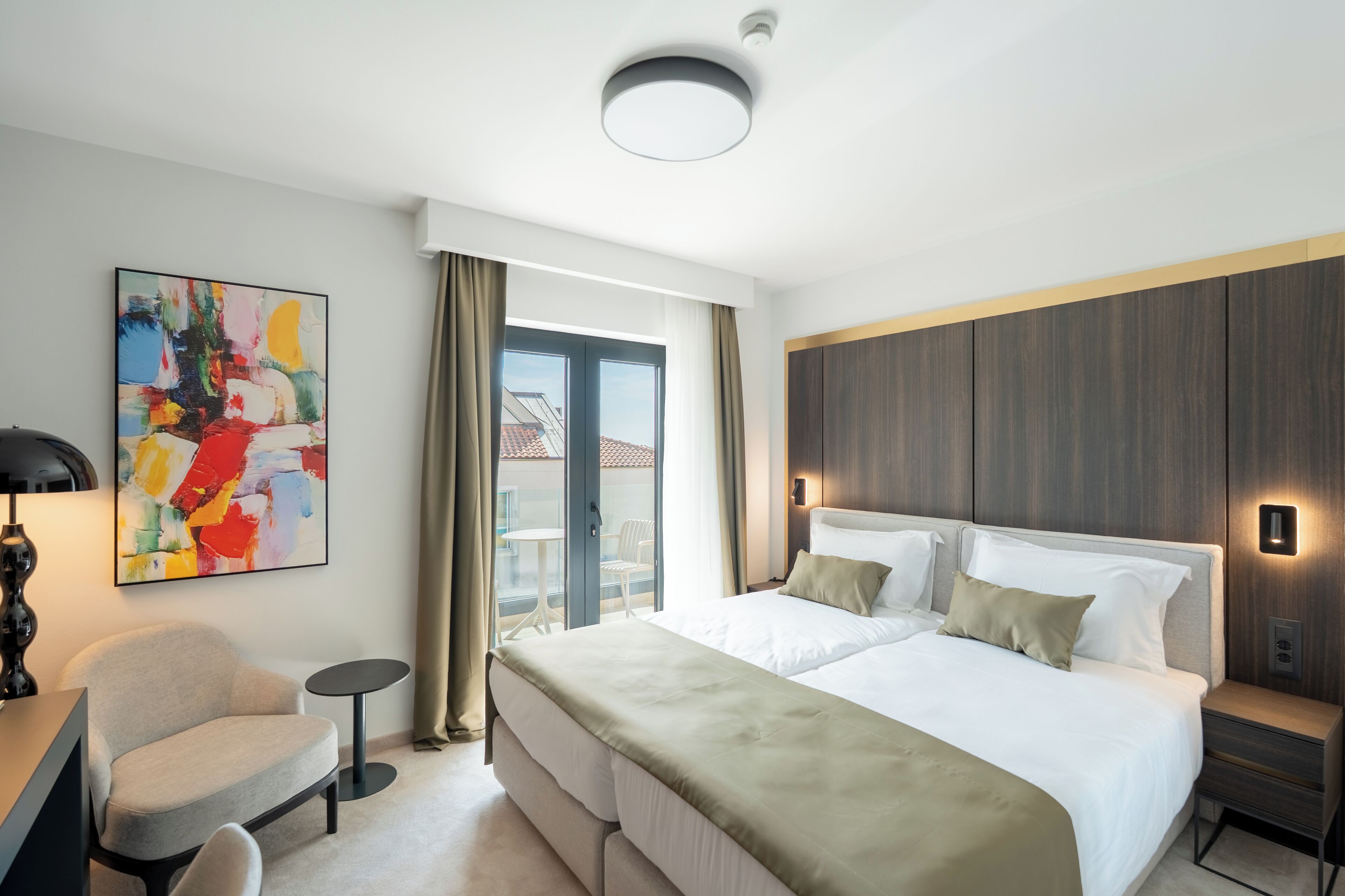 Ett modernt hotellrum med en stor säng, en stol, ett litet bord och en färgstark, abstrakt målning på väggen.